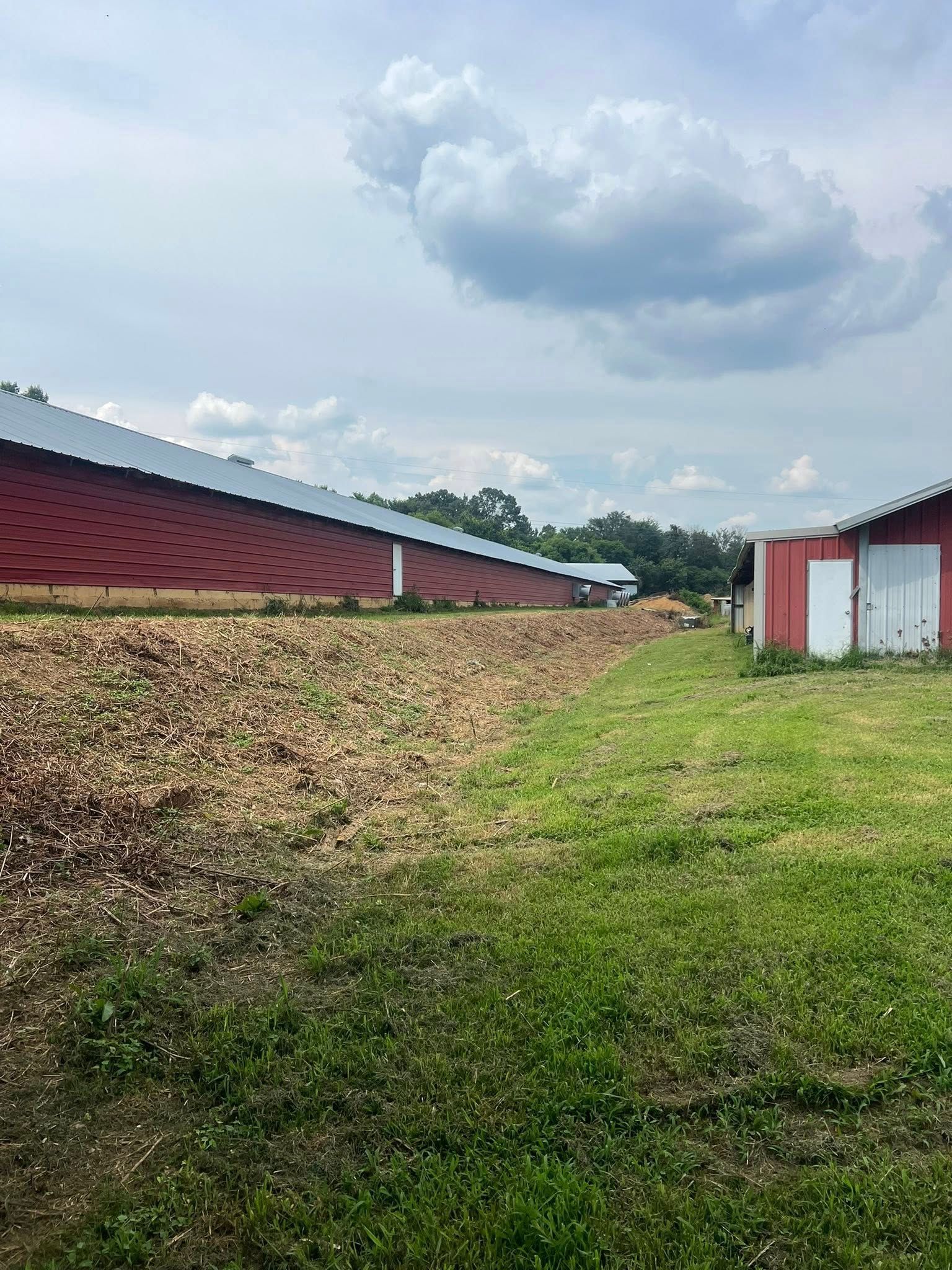 A long red agricultural shed with a mesh side wall sits next to a grassy field under a cloudy blue sky.