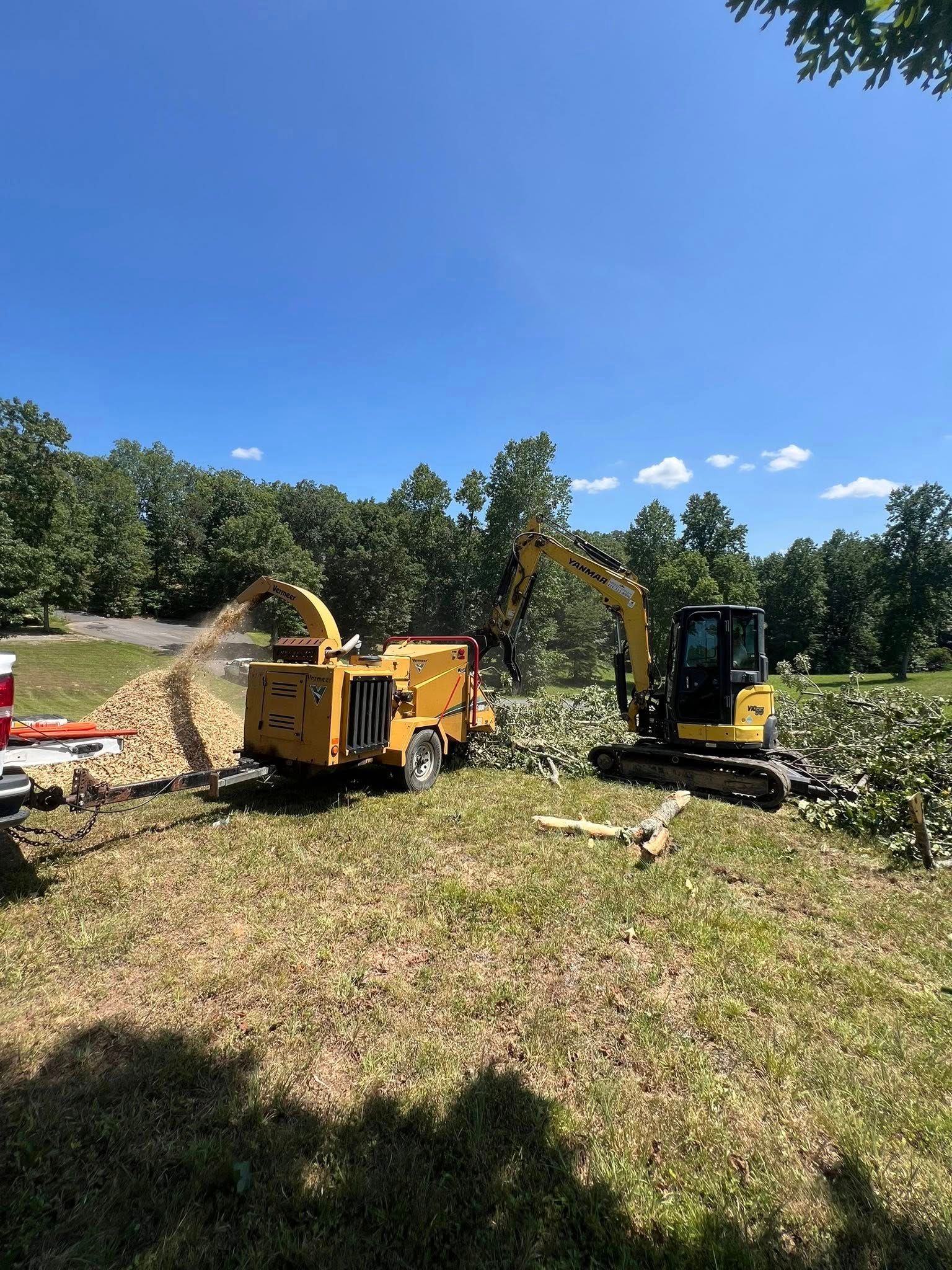 A yellow excavator and wood chipper operating in a grassy field with a forest in the background under a blue sky.