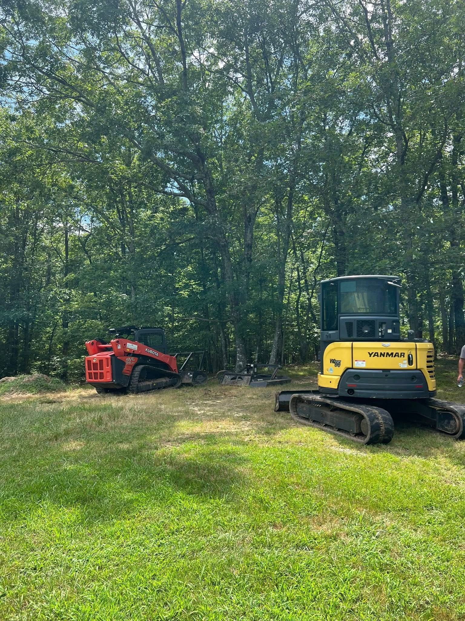 A red skid-steer and a yellow mini-excavator parked on a grassy field in front of a dense forest.