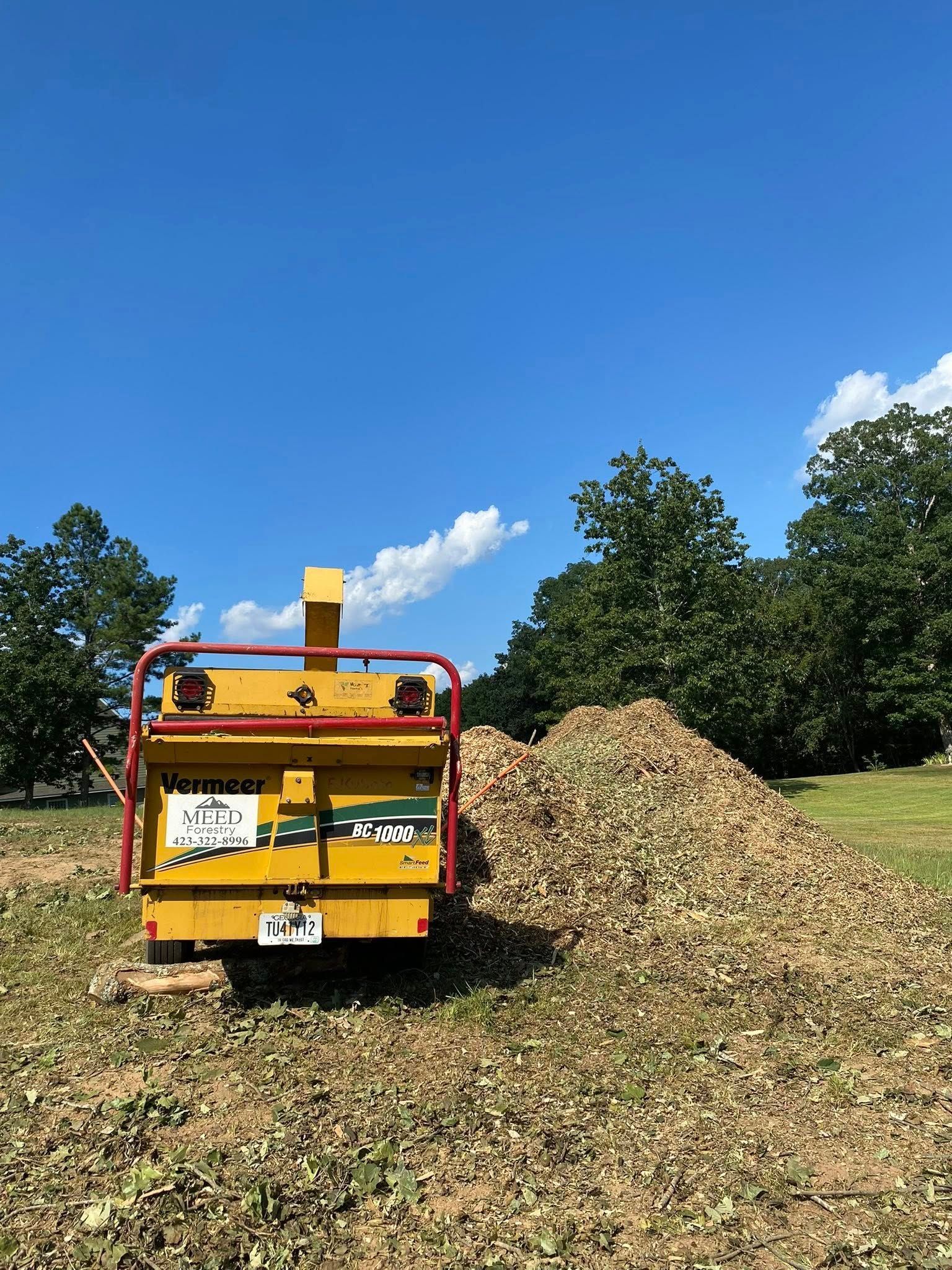 A yellow Vermeer wood chipper stands in a grassy field next to a large pile of wood chips under a clear blue sky.