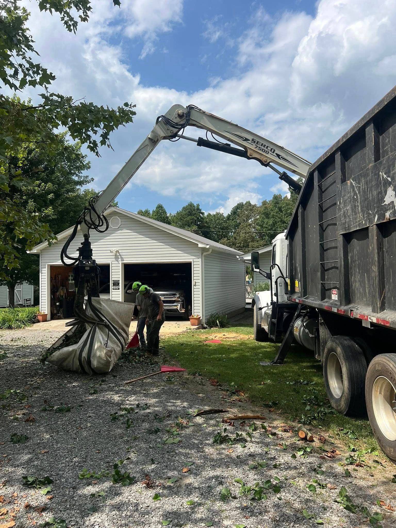 A truck uses a hydraulic grapple arm to lift debris into its large black dumpster next to a house with a garage.