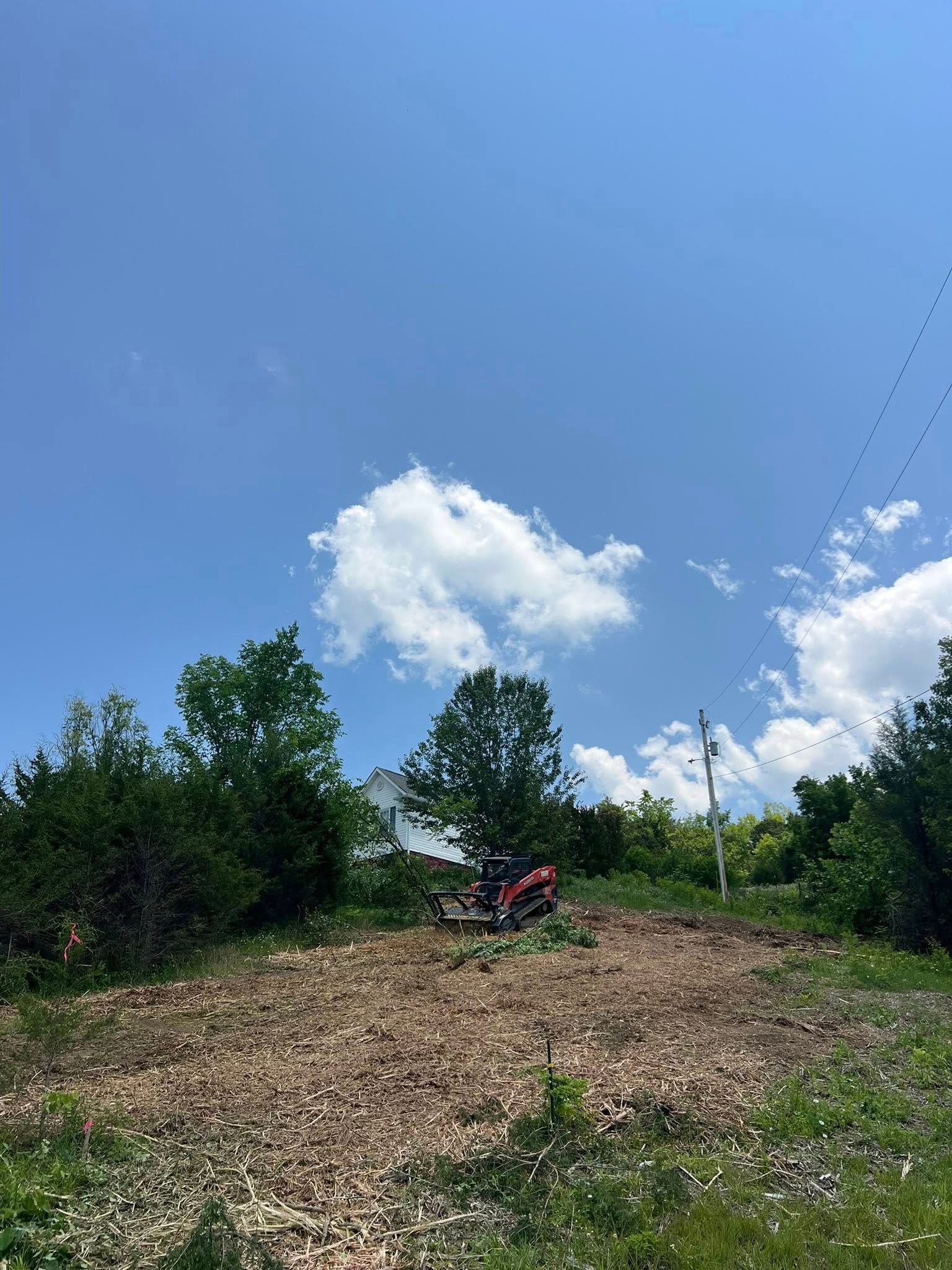 A tractor sits on a cleared, woody patch of land beneath a bright blue sky with scattered white clouds.