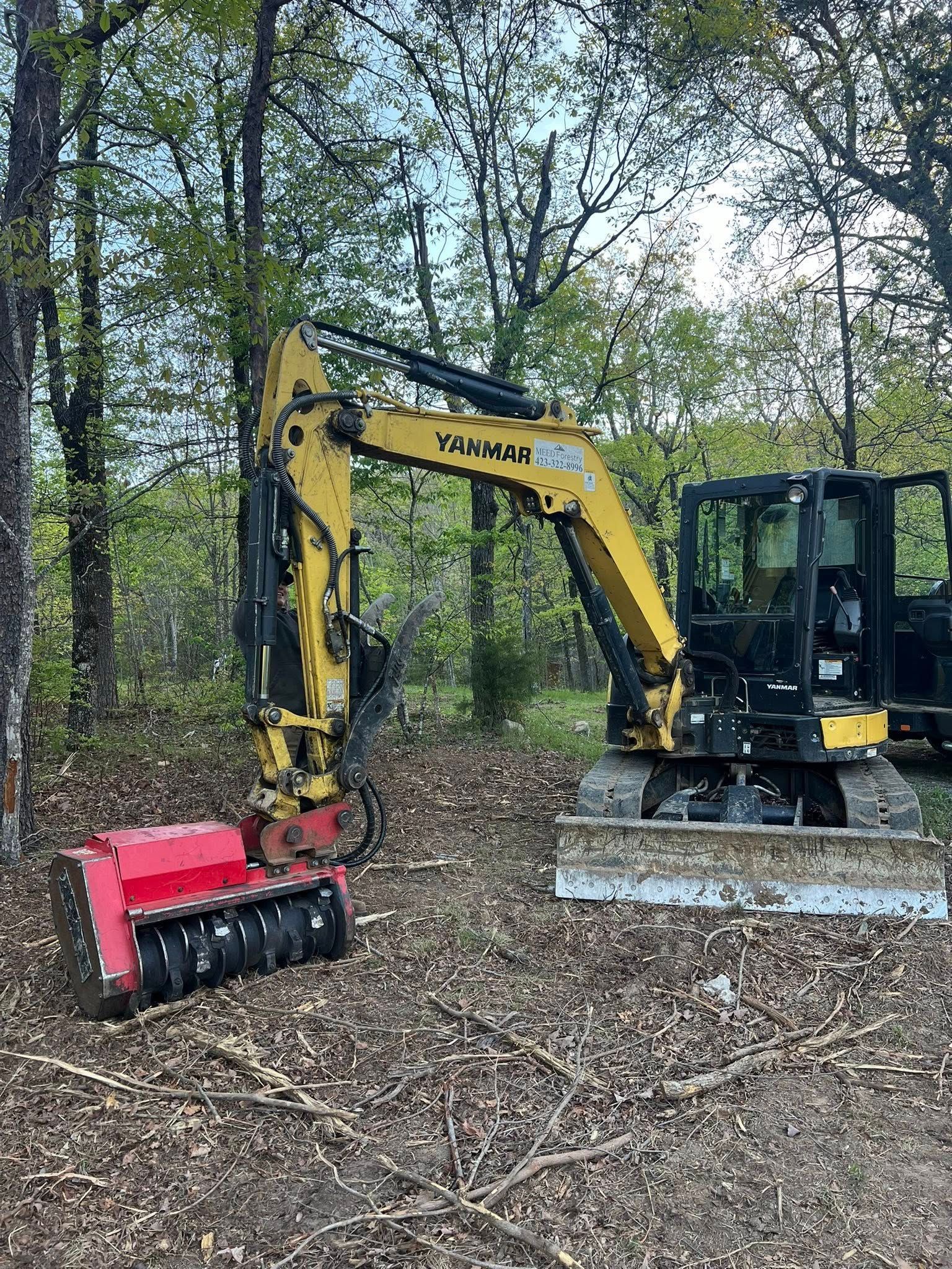 A yellow Yanmar excavator equipped with a red mulcher attachment parked in a wooded area.