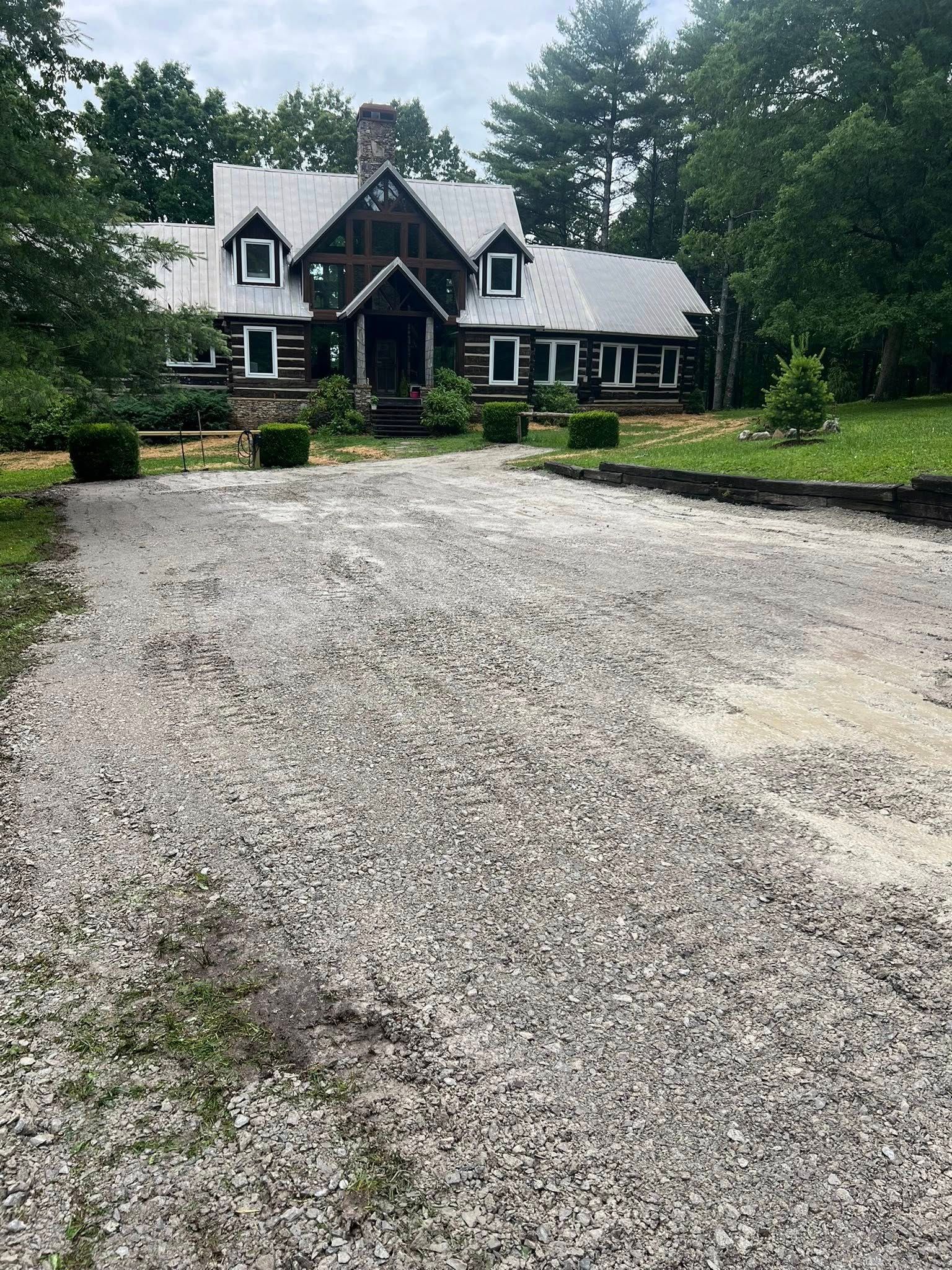 A gravel driveway leads to a two-story log house with a metal roof and gabled windows surrounded by lush green trees.