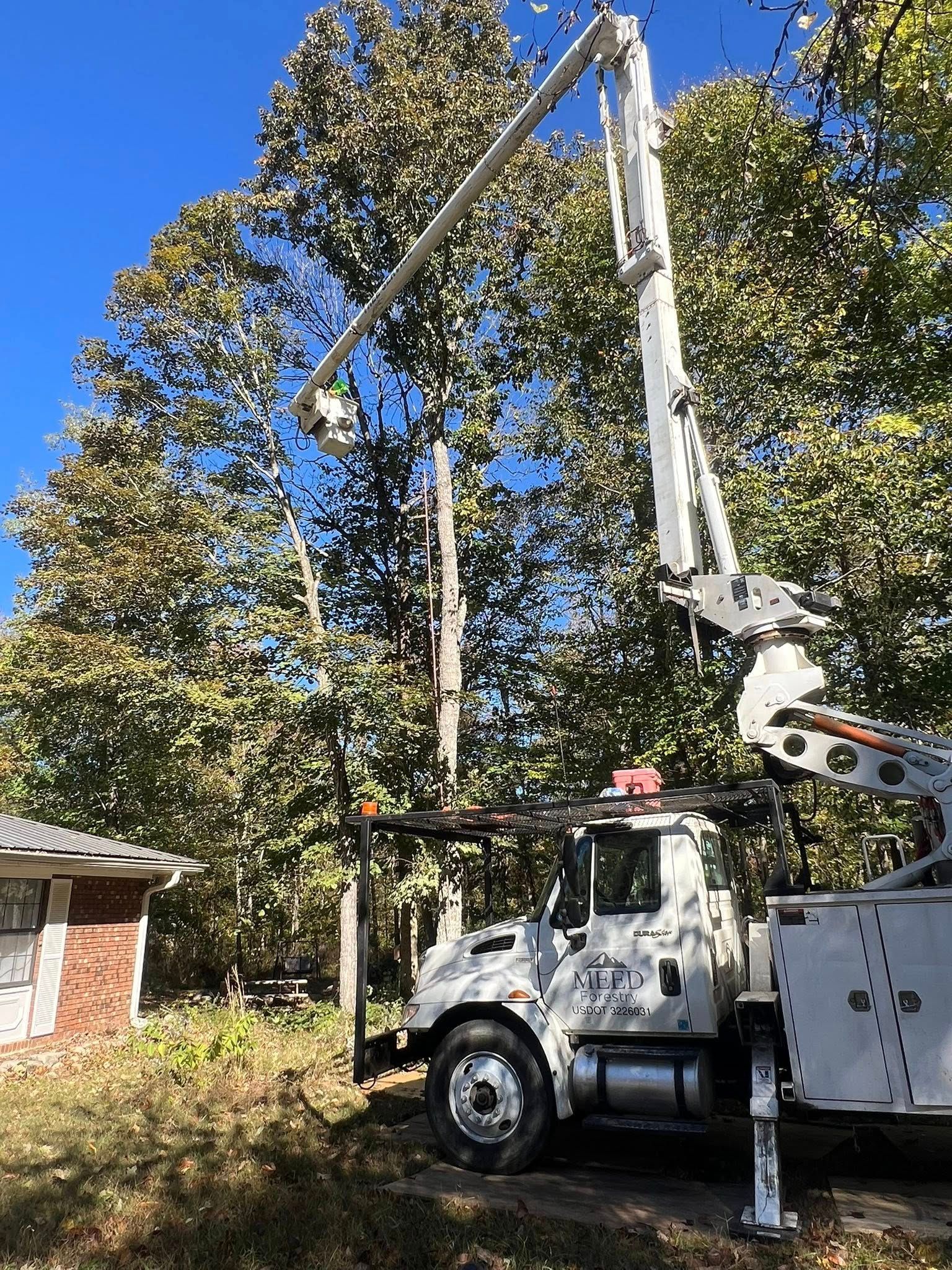 A white utility truck with an extended boom lift sits in a yard next to a brick house and tall trees on a sunny day.