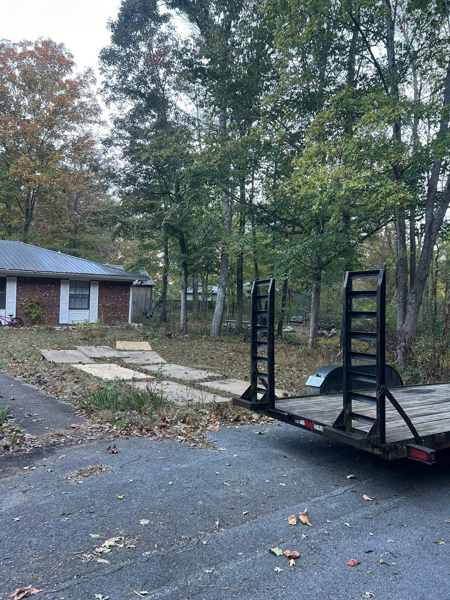 A flatbed utility trailer with loading ramps sits on a gravel driveway in front of a brick house surrounded by trees.