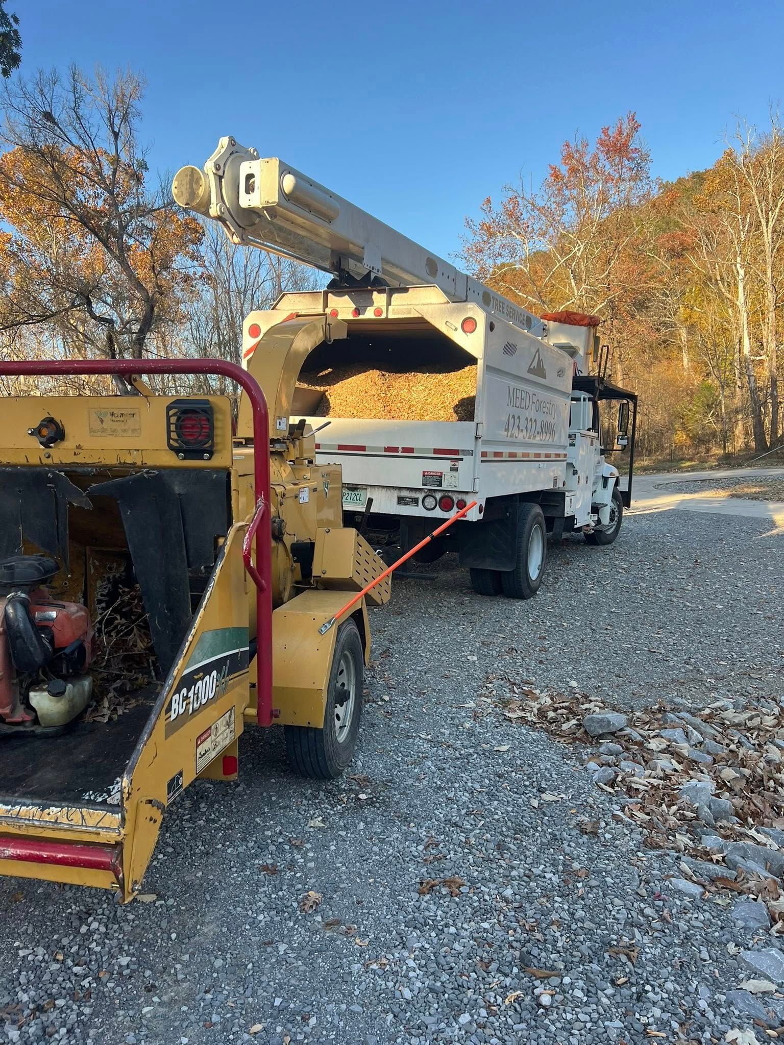 A wood chipper attached to a utility truck filled with wood chips, parked on a gravel surface near trees under a blue sky.
