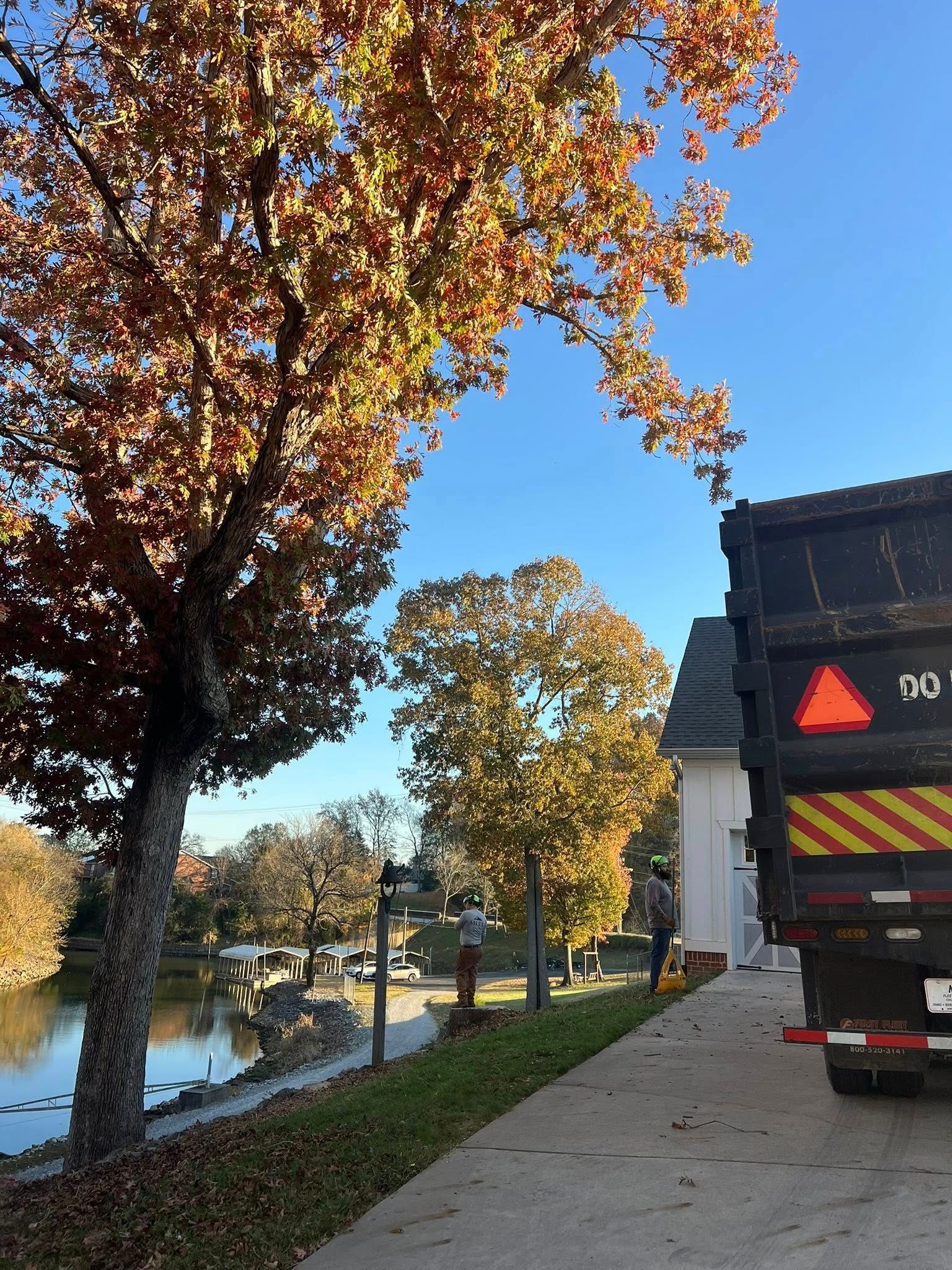 A truck parked on a driveway near a house and a small pond, framed by trees with autumn-colored foliage under a blue sky.