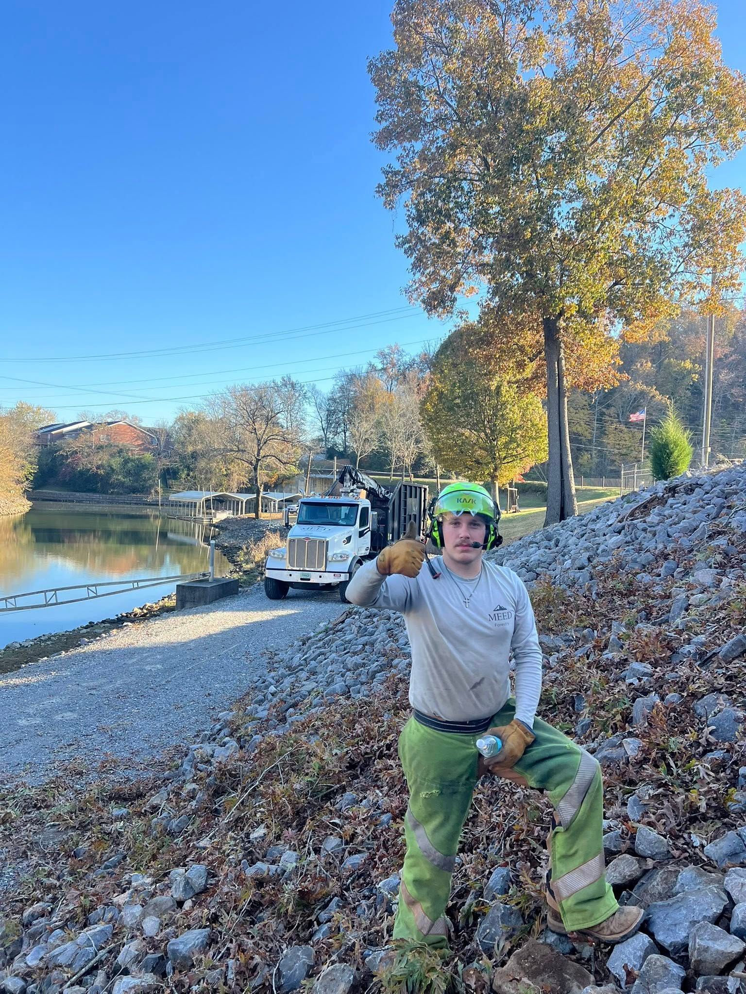 A worker in a helmet, safety gear, and work pants stands on a rocky, leaf-covered embankment with a truck by a pond.