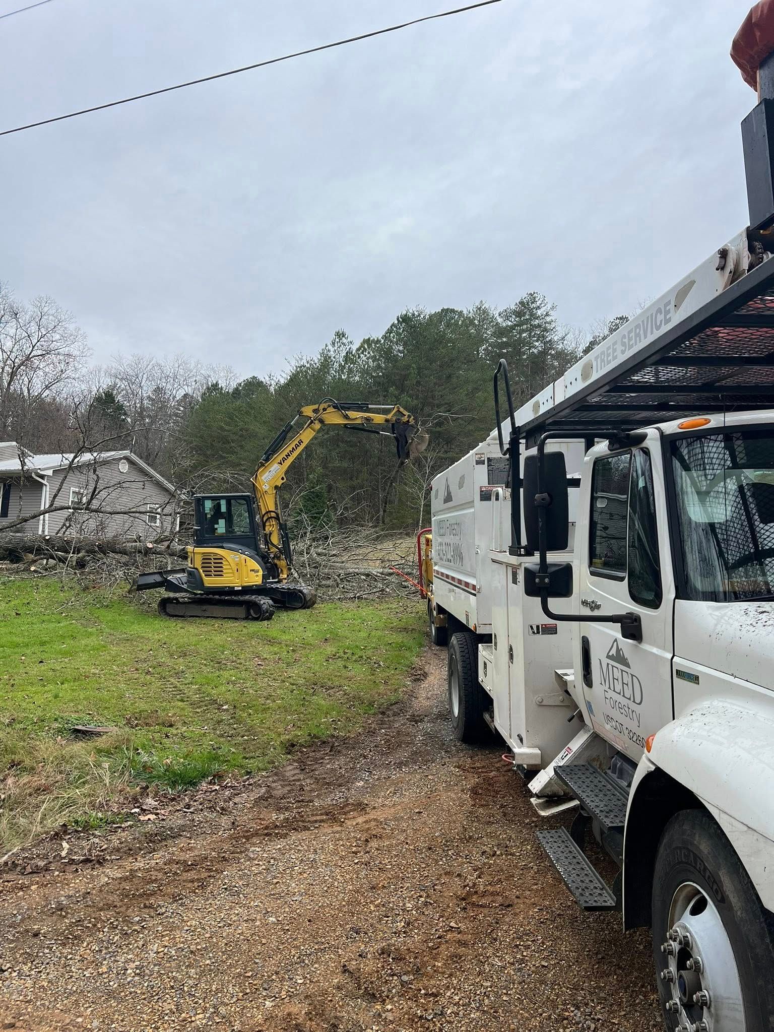 A yellow mini excavator works in a yard next to a white utility truck on a gravel path near a house and trees.