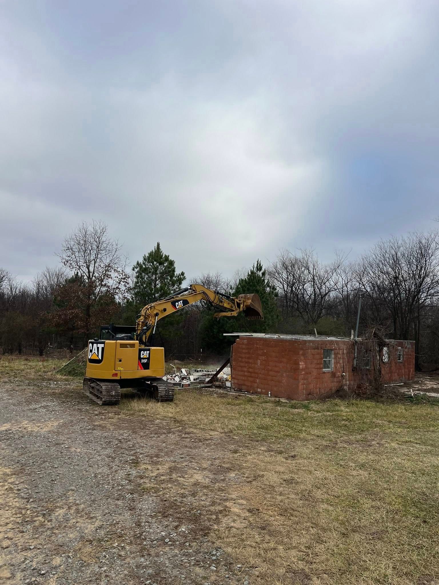 A yellow excavator demolishes a small red brick building in an open, grassy field under a cloudy sky.