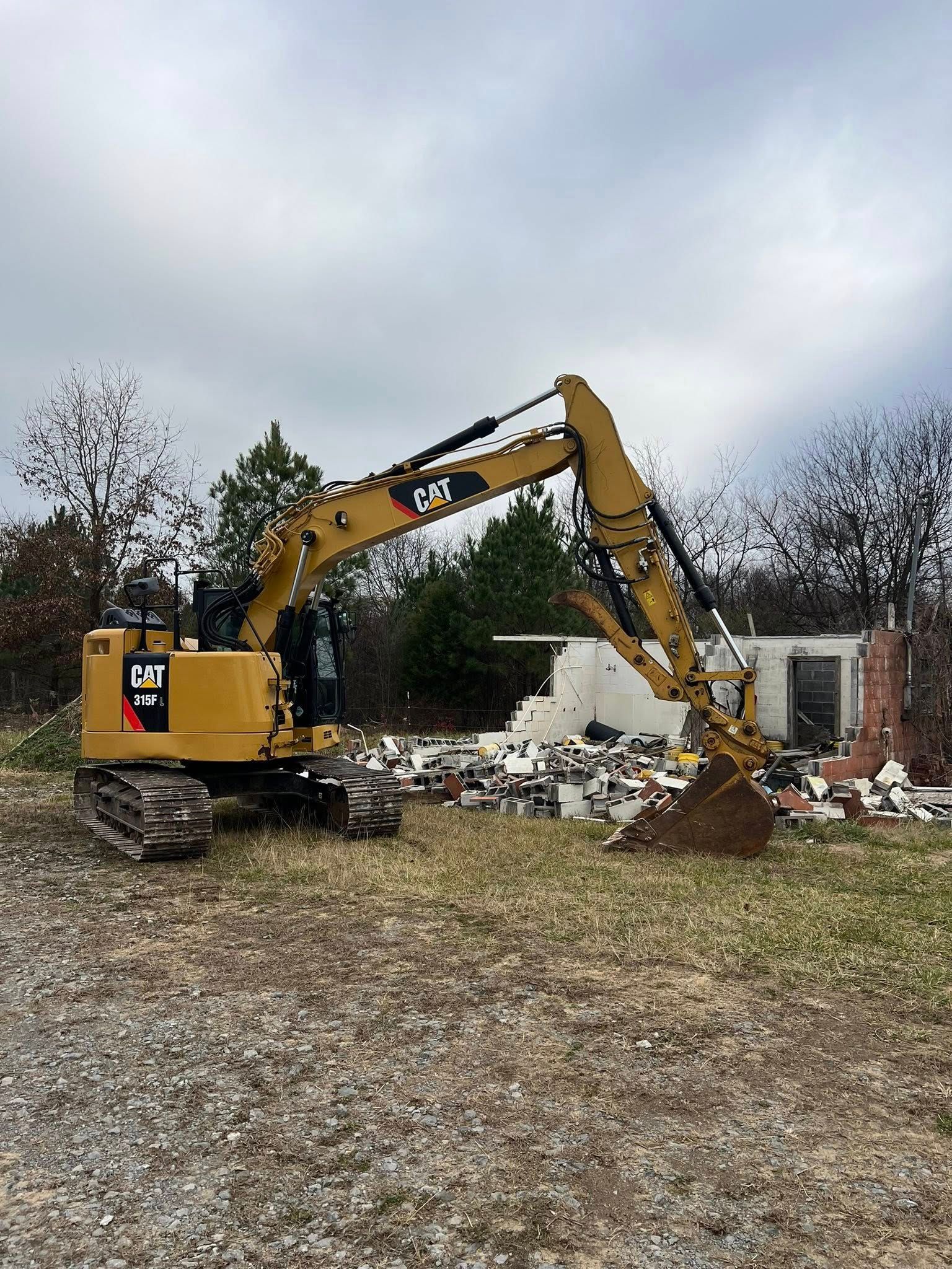 A yellow hydraulic excavator stands in a grassy lot near the rubble of a demolished building under a cloudy sky.