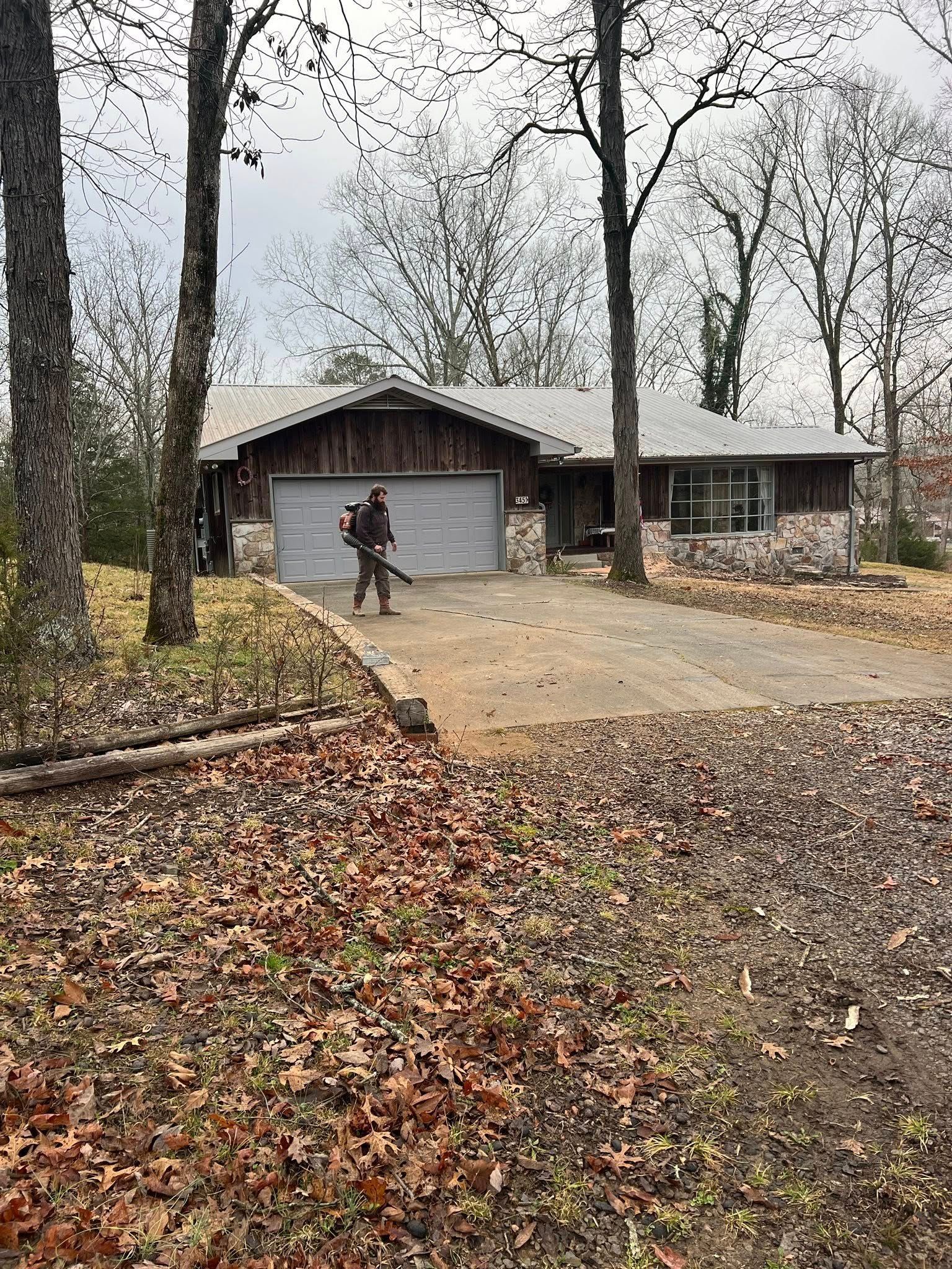 A person uses a leaf blower to clear leaves from a driveway in front of a house with stone accents and a metal roof.