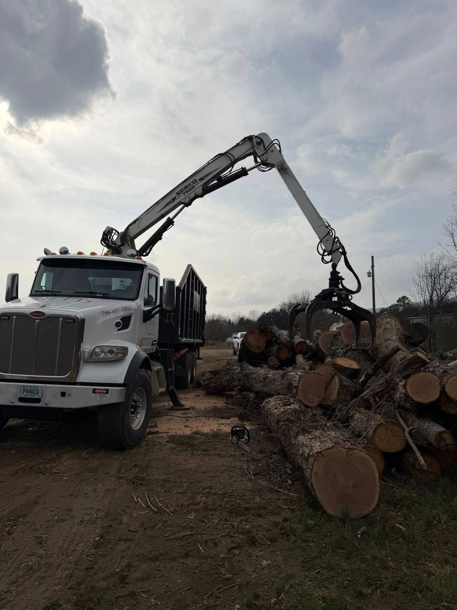 A white boom truck with a log grapple claw lifting wood from a pile in a rural, outdoor setting.