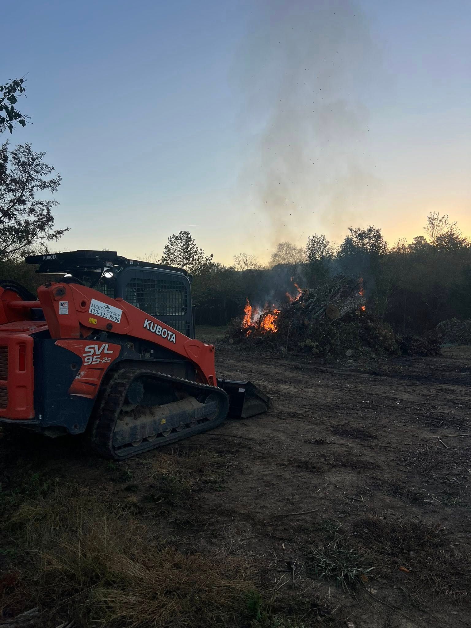 A red skid-steer loader sits in a field near a burning pile of brush under a sunset sky.