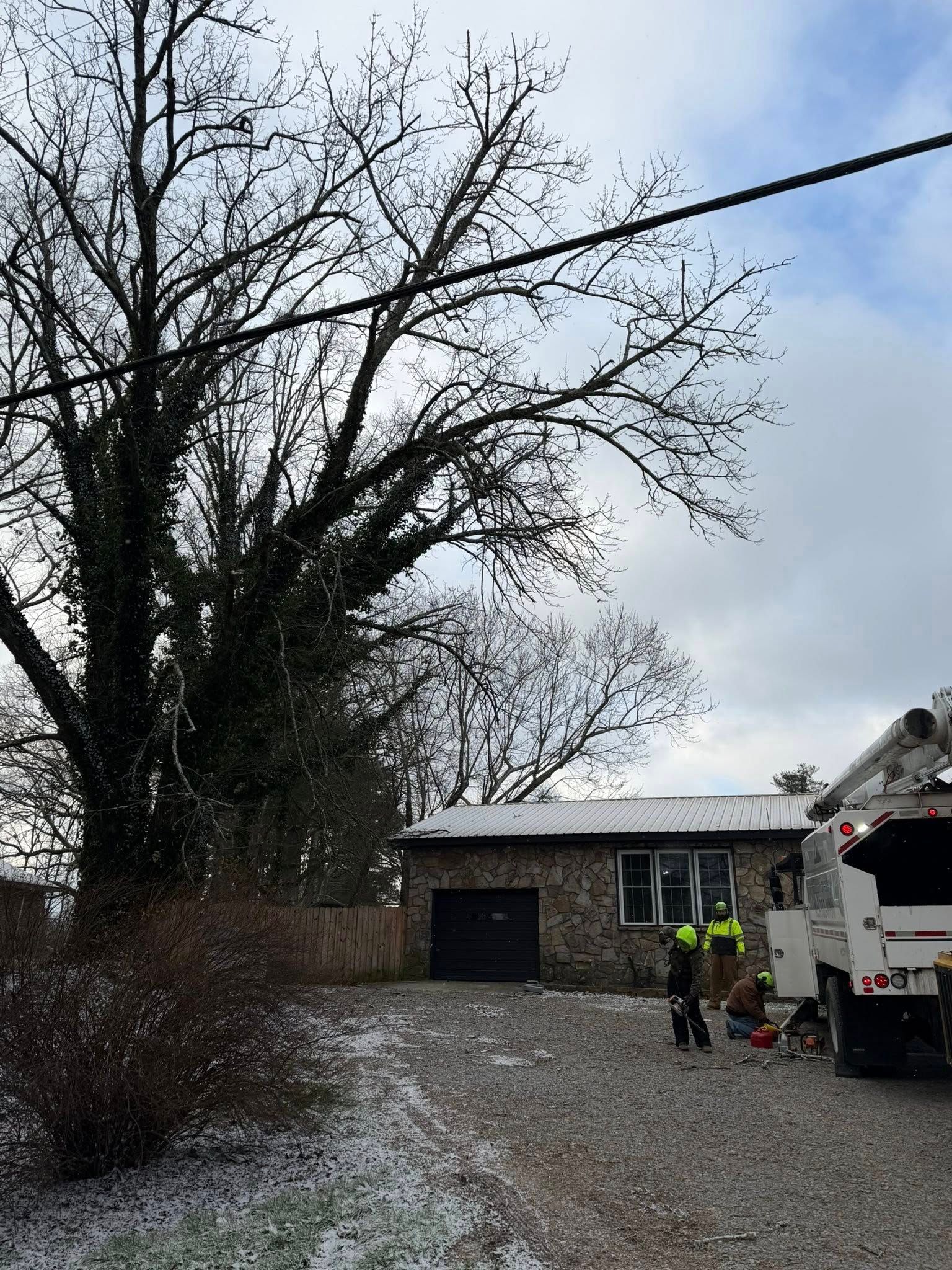 A utility truck is parked on a gravel driveway near a stone house and a large tree under a cloudy, light-snow sky.