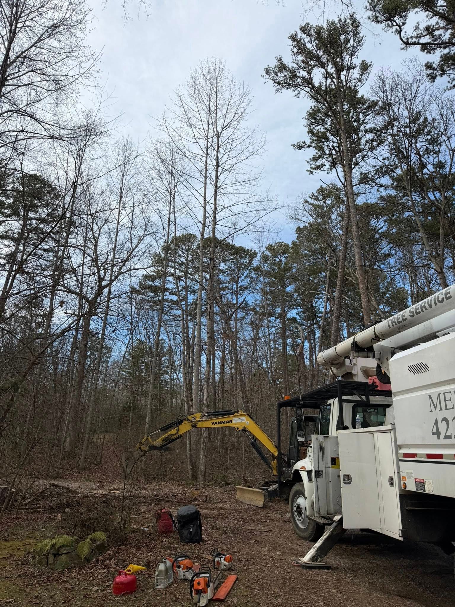 A white utility truck with an extended boom sits in a wooded area with yellow construction equipment and scattered tools.