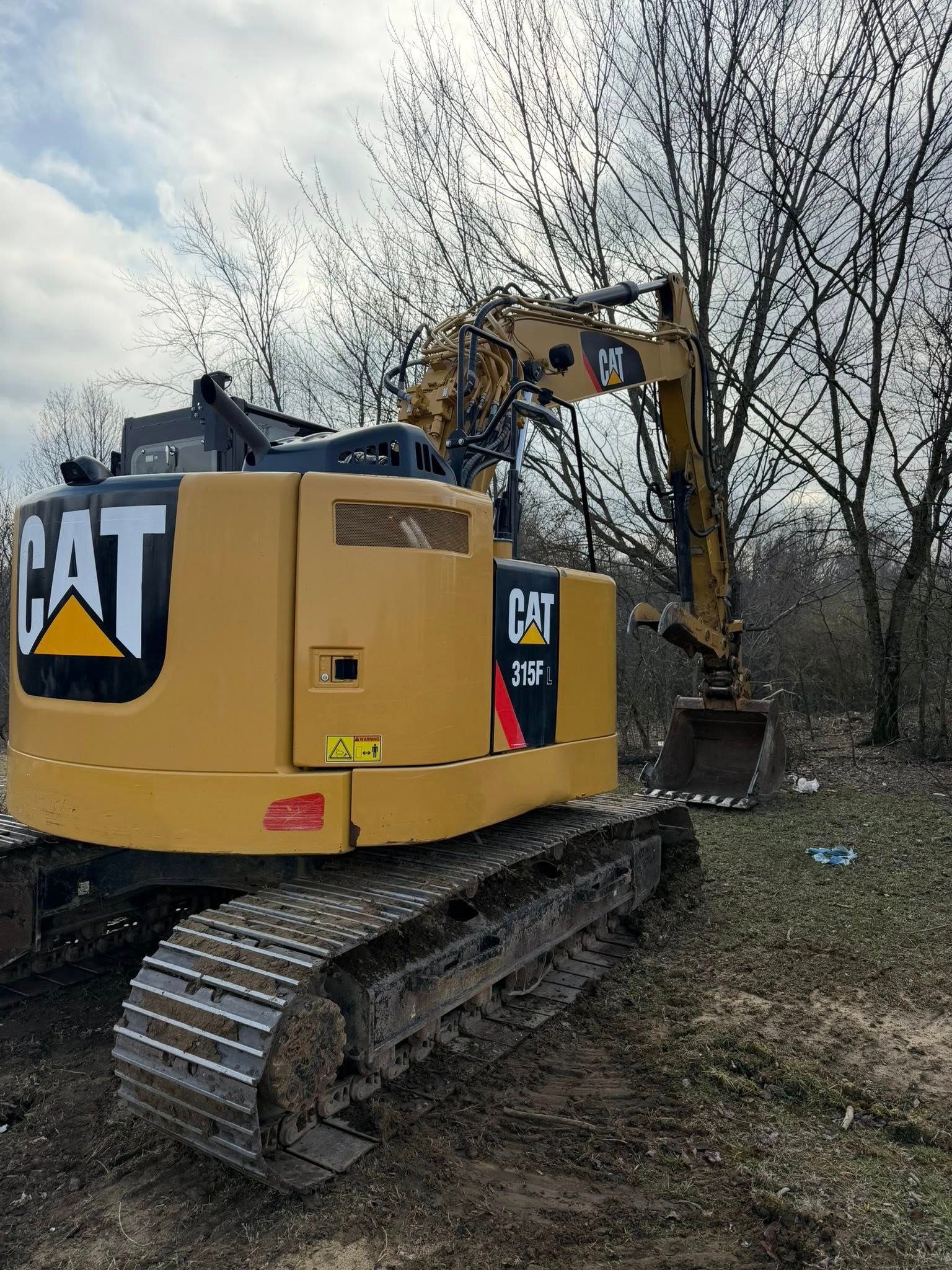 A yellow Caterpillar excavator parked on a dirt field with a bucket resting on the ground near bare trees.