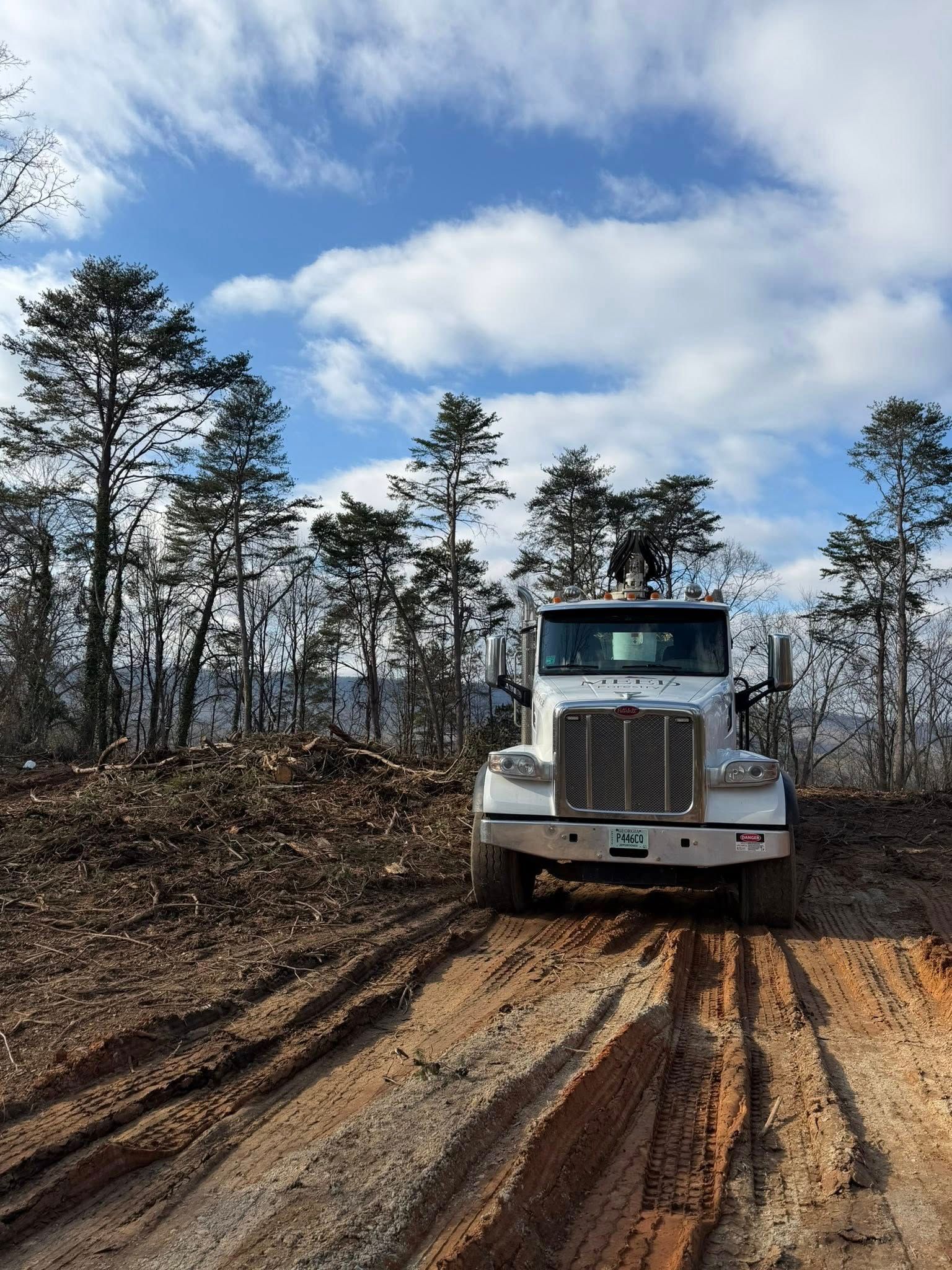 A white semi-truck parked on a muddy dirt road in a wooded area with cleared brush under a blue sky.