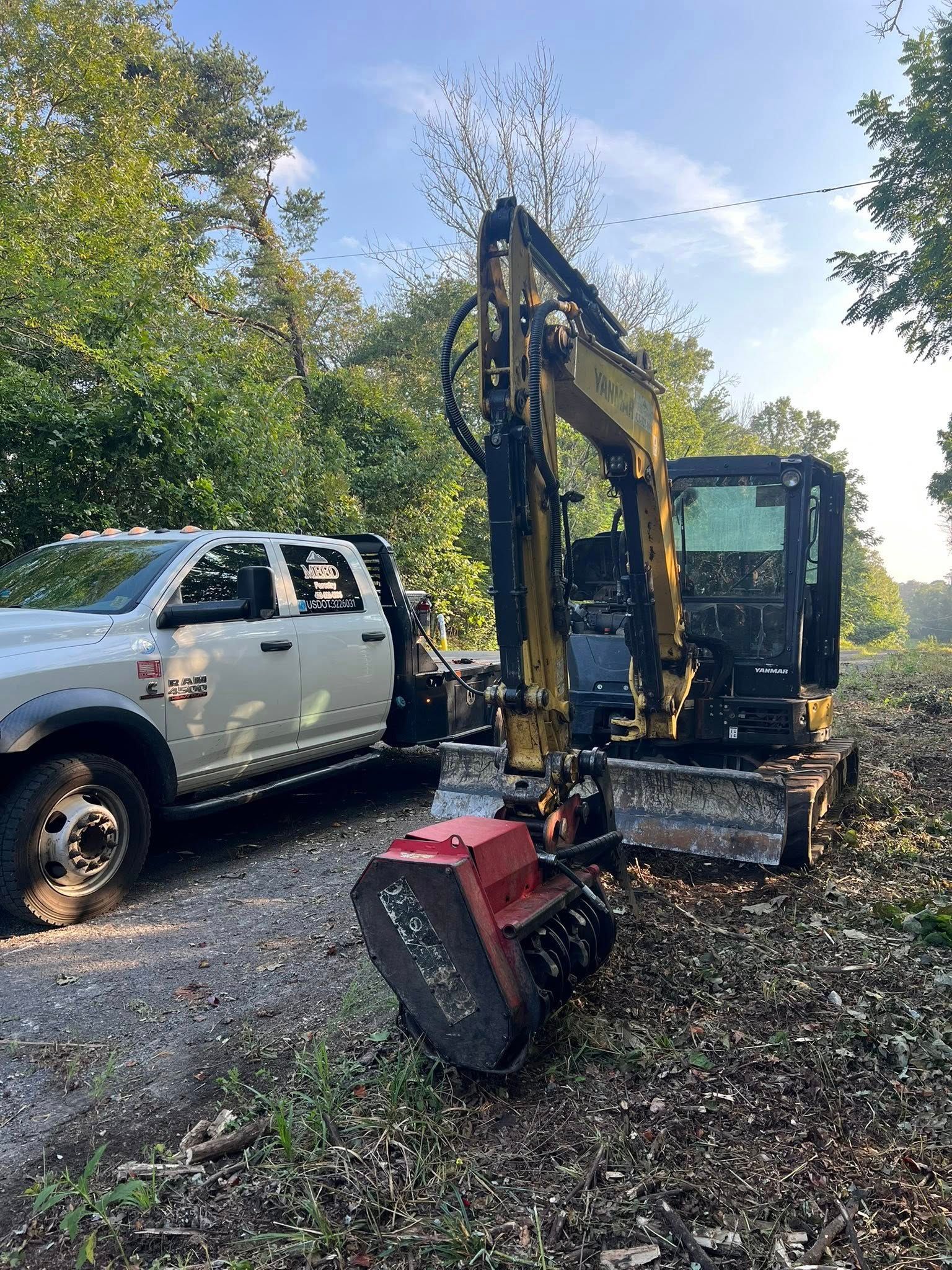 A silver pickup truck parked next to a yellow excavator equipped with a red mulcher head on a gravel path near trees.