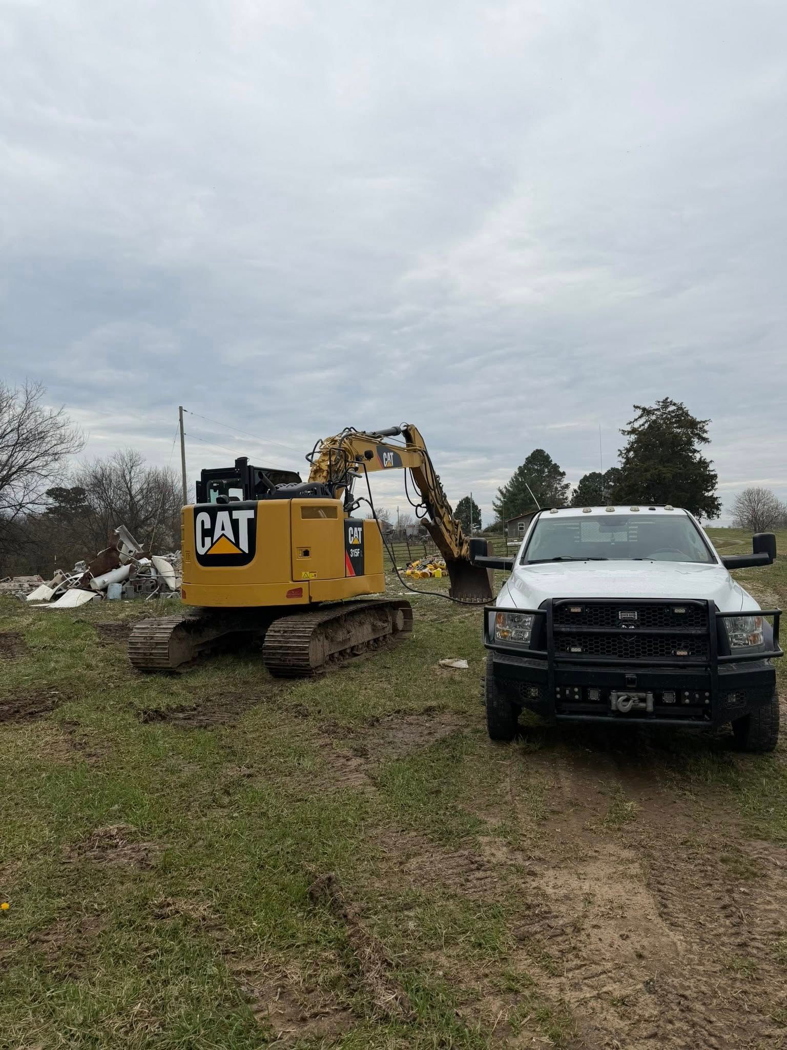 A yellow Caterpillar excavator parked next to a white pickup truck in an open, grassy field under a cloudy sky.