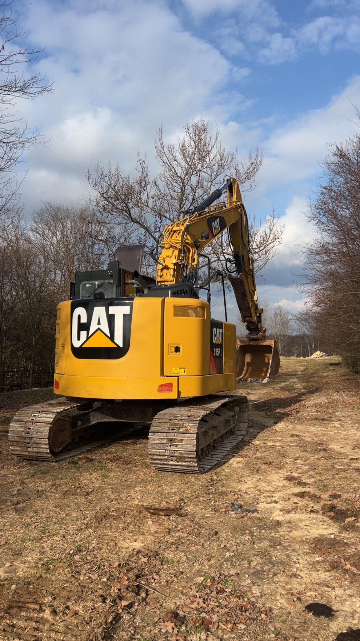 A yellow CAT excavator parked on a field with bare trees under a cloudy blue sky.