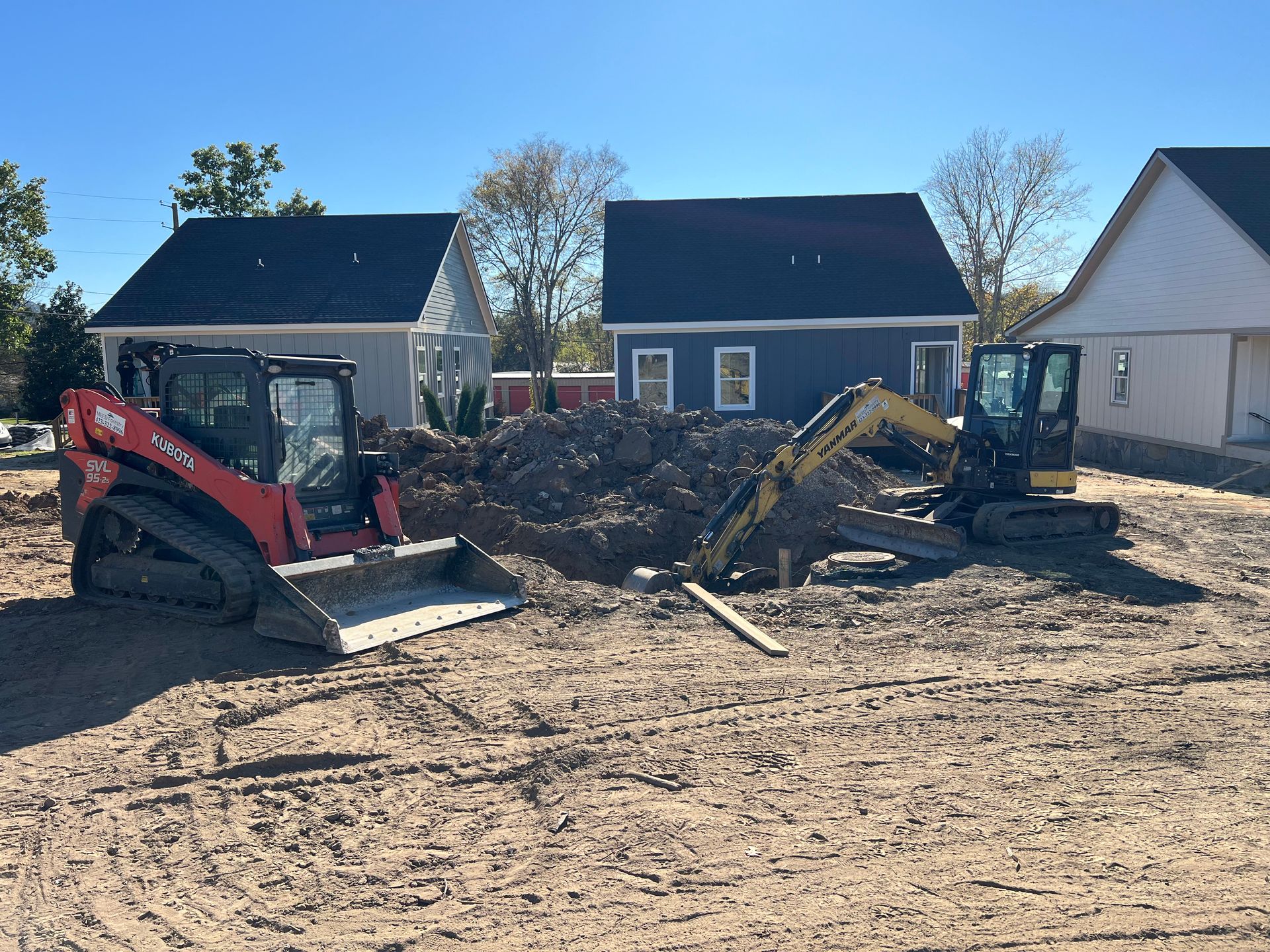 A red skid steer and a yellow mini excavator sit on a dirt construction site in front of two new houses.