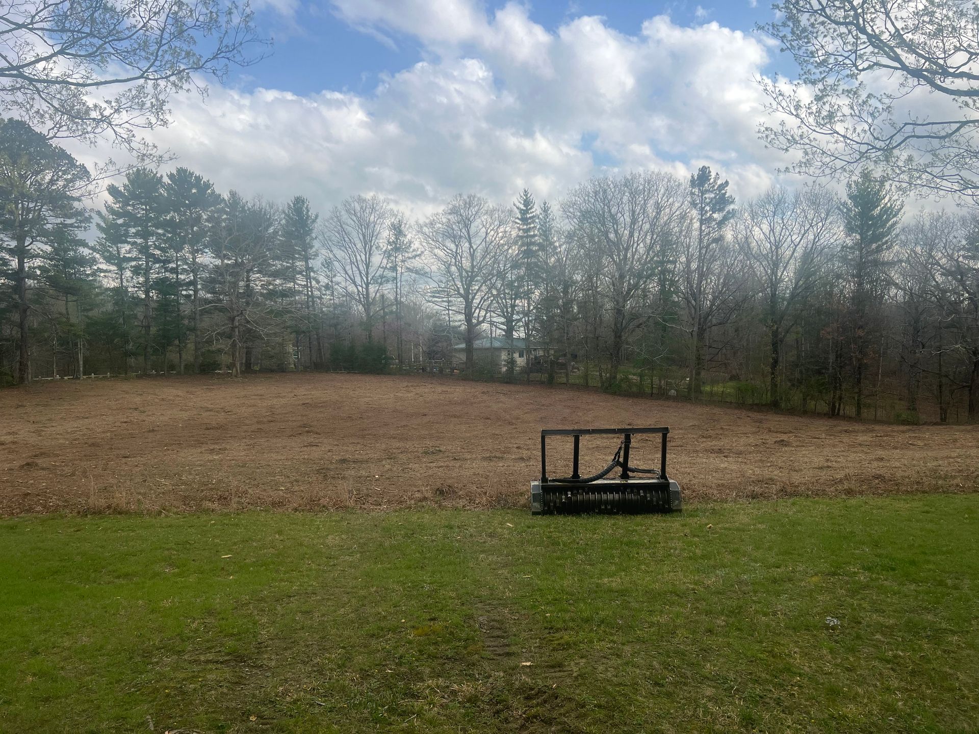 A brush hog mower sits in a large, dormant field edged by a dense forest under a cloudy blue sky.