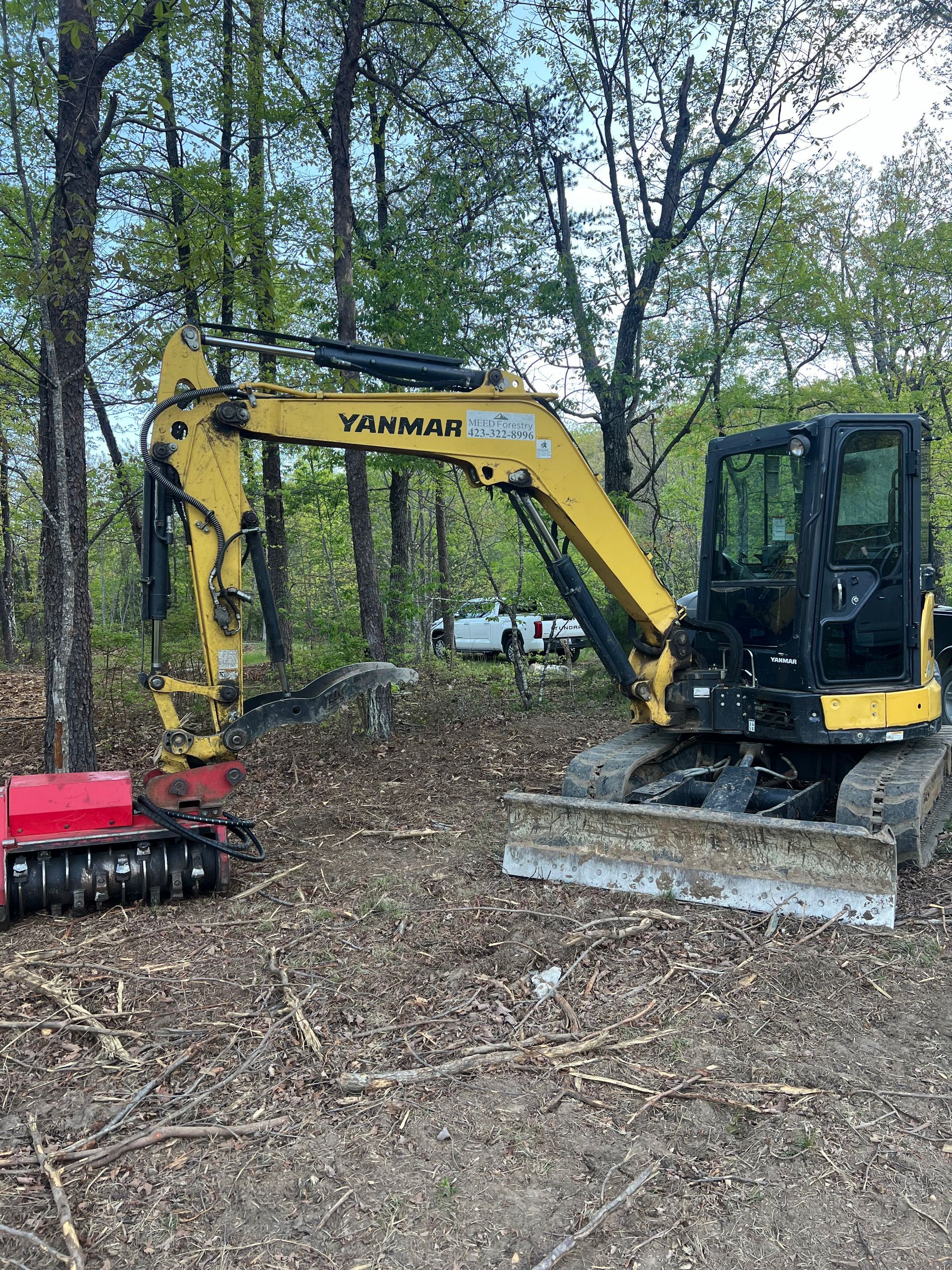 A yellow Yanmar excavator with a red mulcher attachment parked in a wooded area.