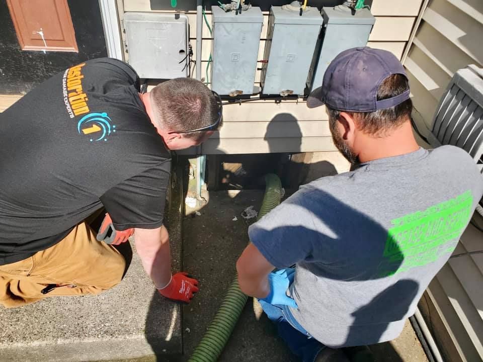 Two workers in work shirts and gloves kneel on a concrete porch, working with a long green hose near utility boxes.