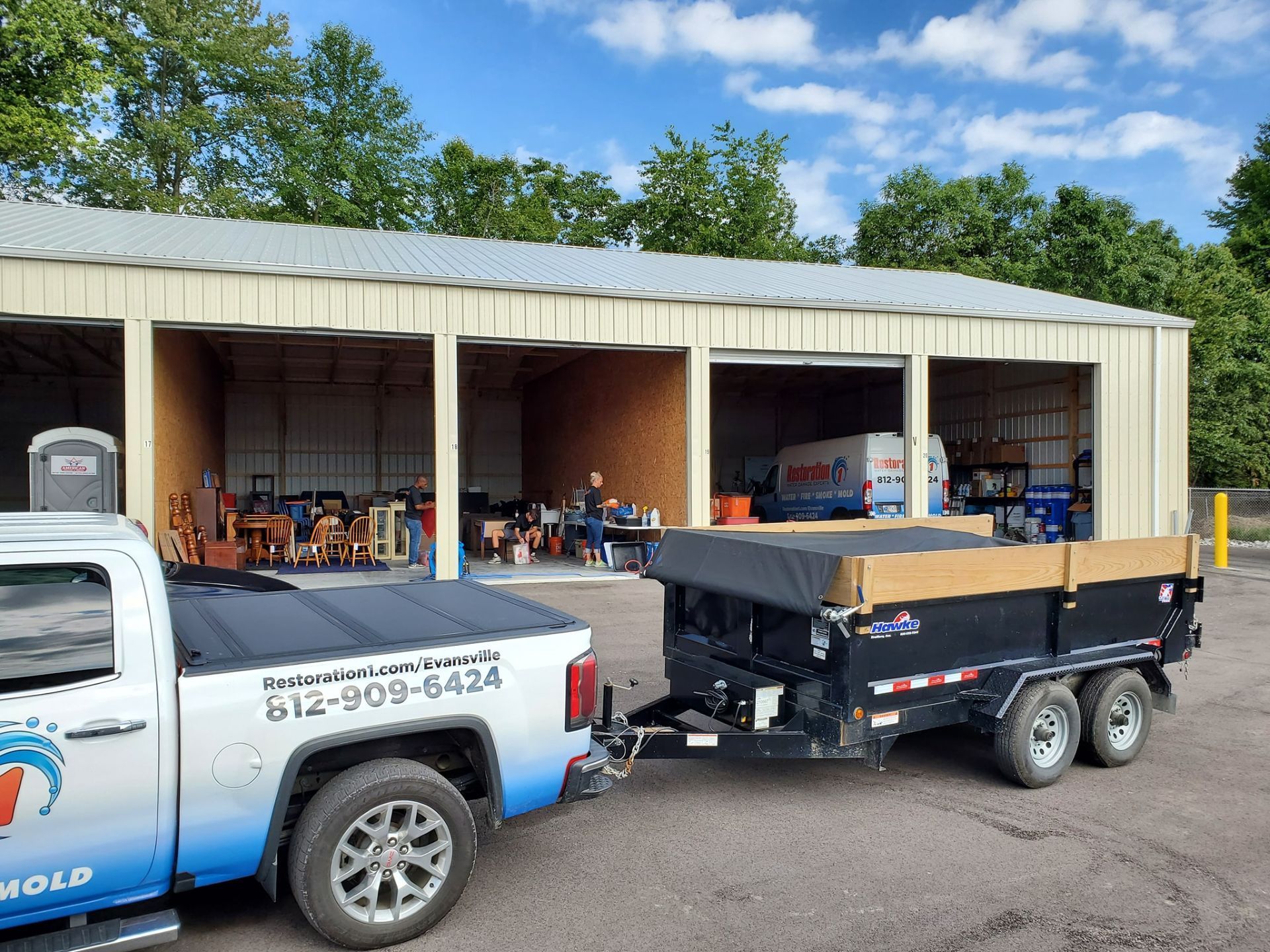 A work truck with a dump trailer parked in front of an open storage bay building on a sunny day.