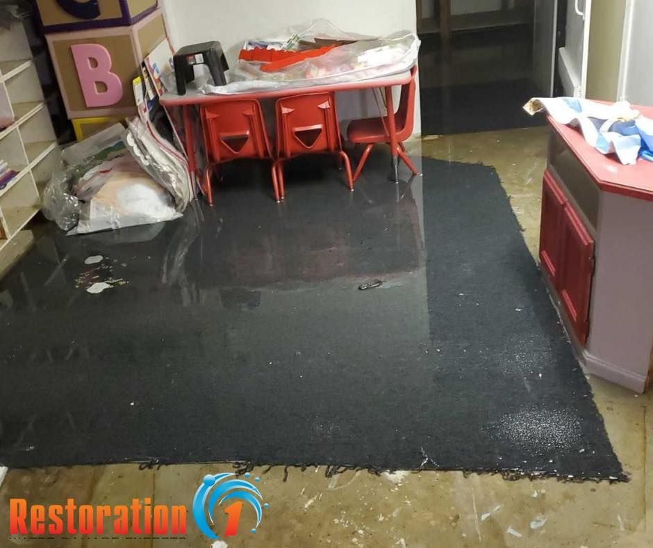 A classroom with a dark, water-damaged rug flooded with standing water, surrounded by red chairs and tables.