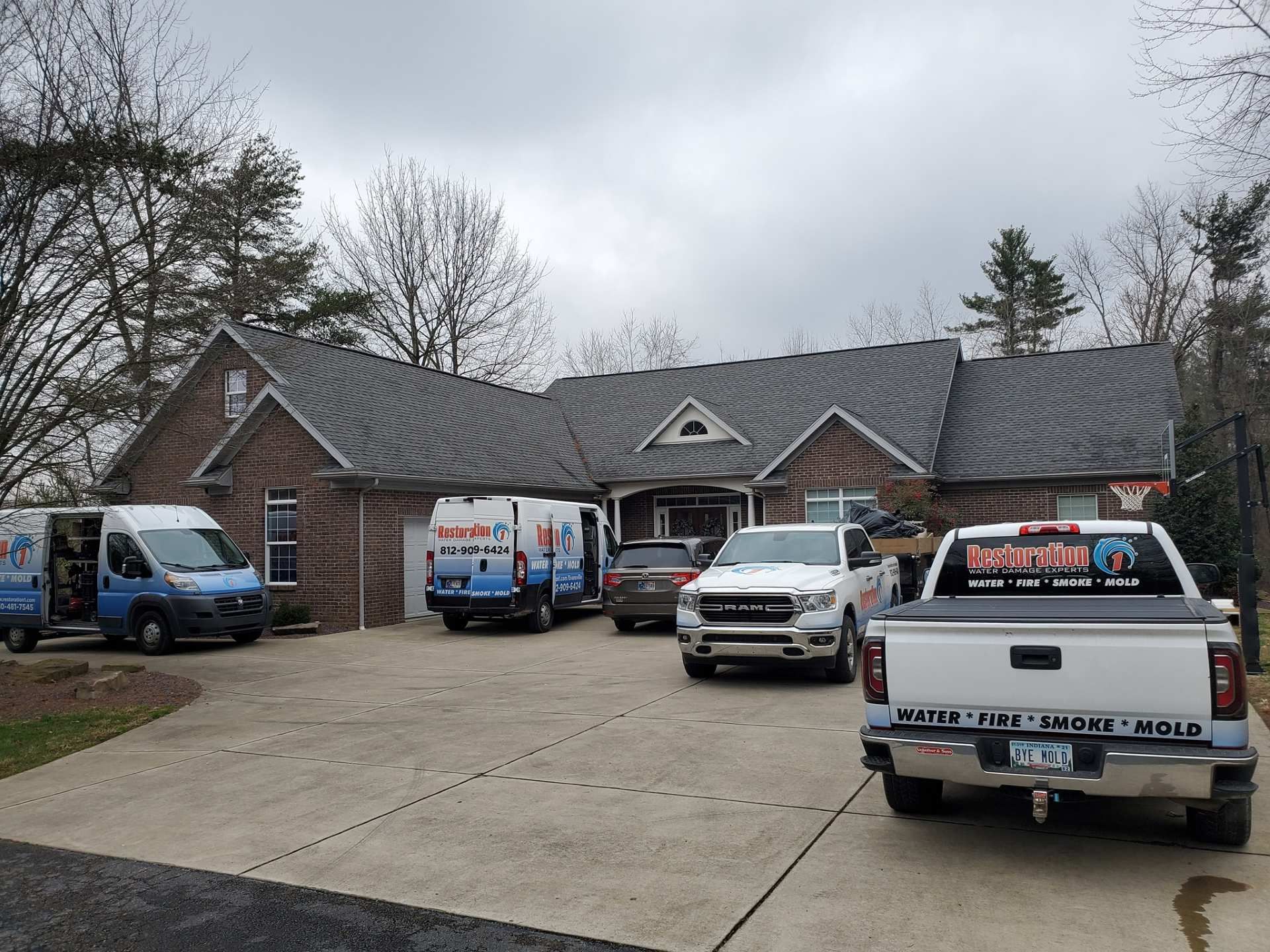 Several work vans and trucks parked in the driveway of a brick house on a cloudy day.