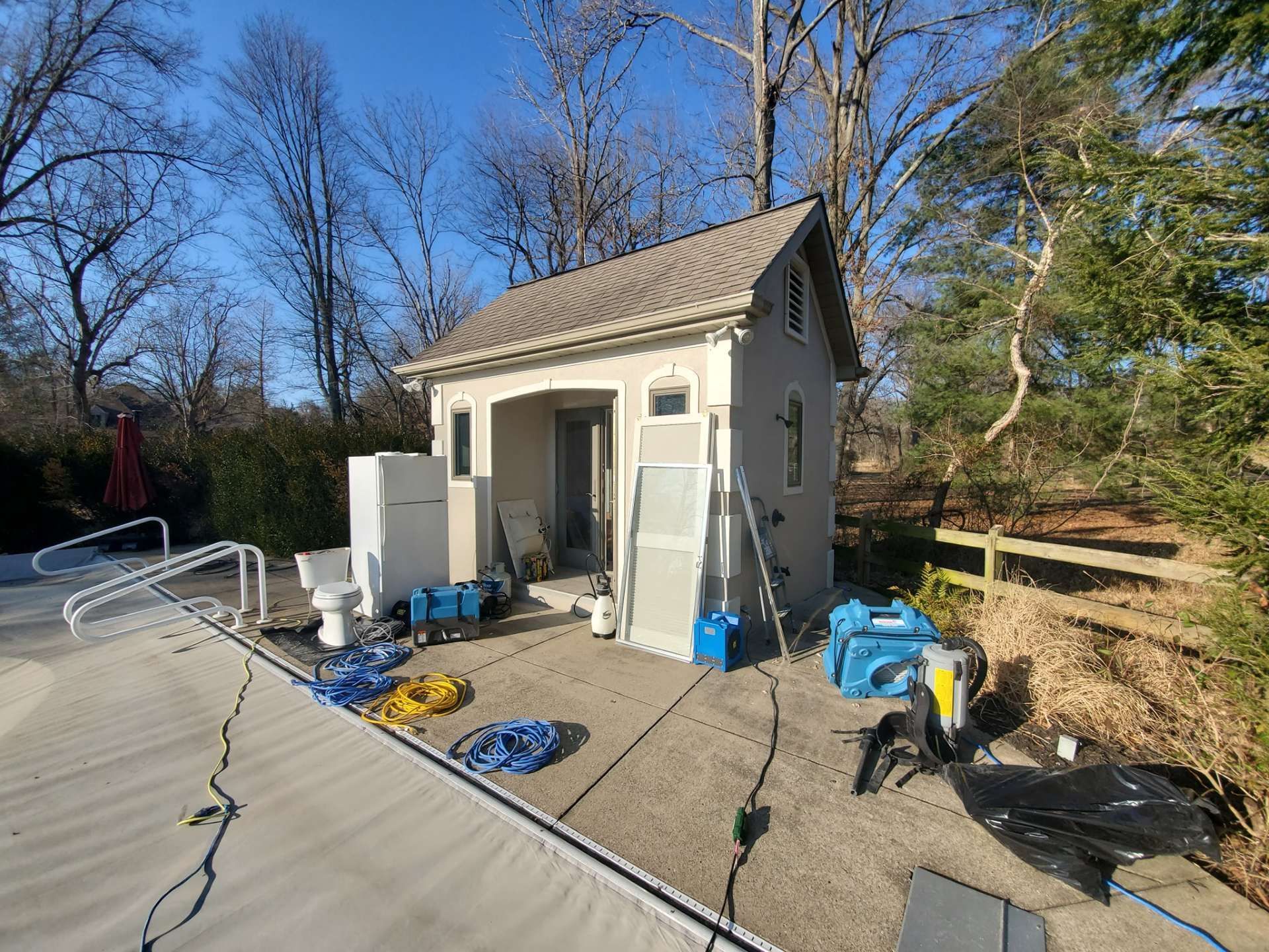A small cream-colored pool house with cleaning equipment, a toilet, and construction supplies on the surrounding patio.