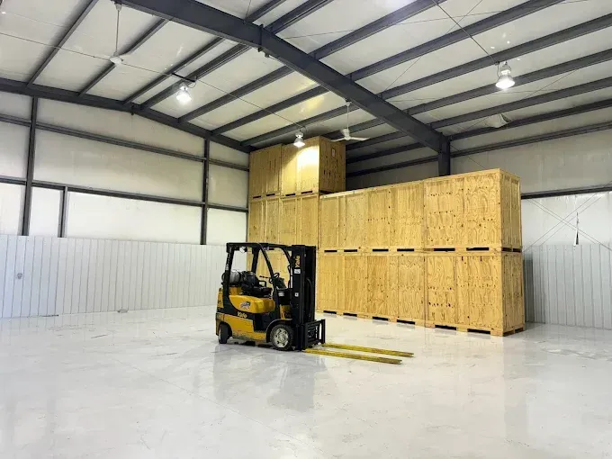 A yellow forklift parked in a spacious, white-walled warehouse next to tall stacks of wooden shipping crates.