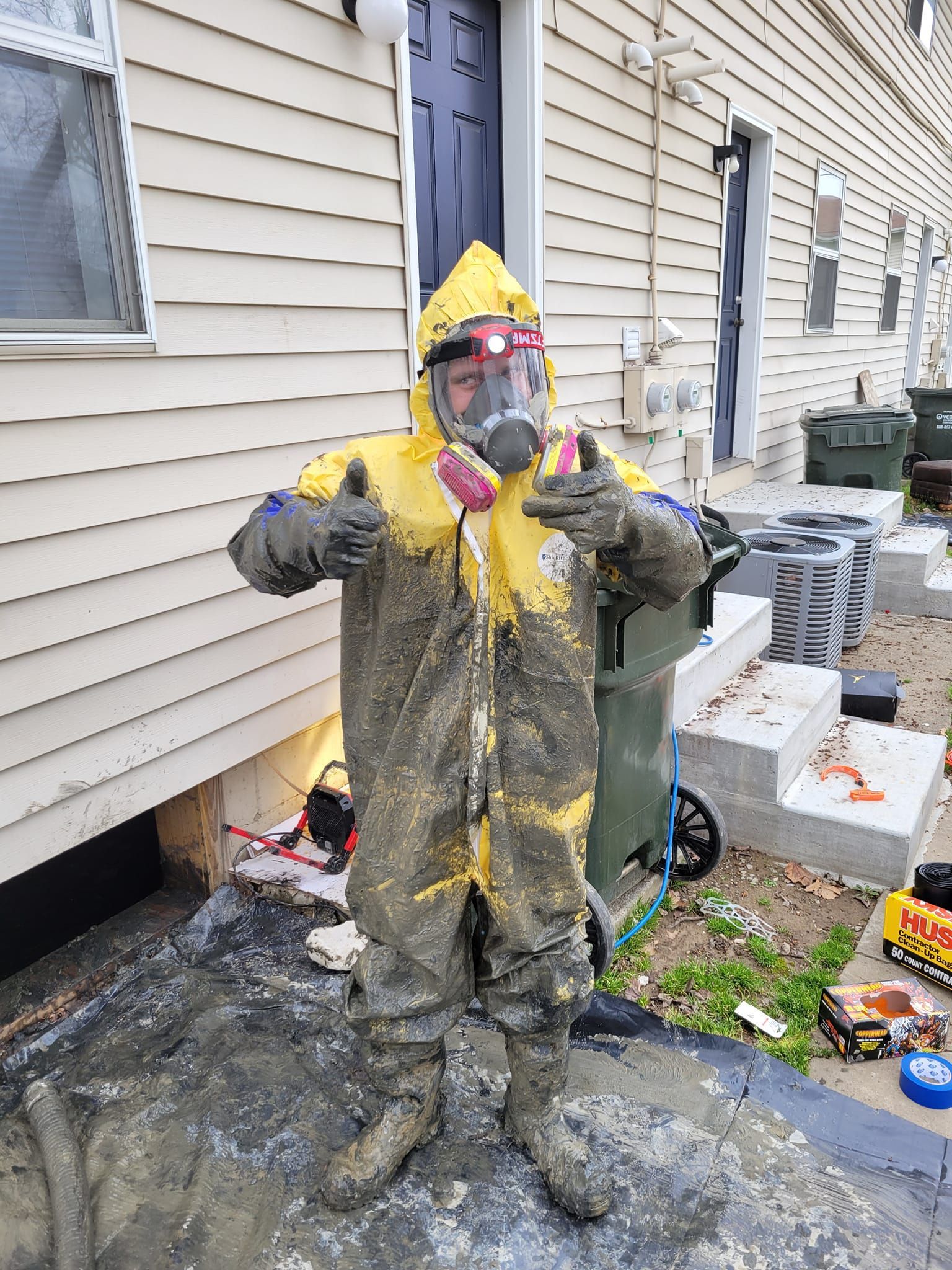 A person in a yellow and mud-caked hazmat suit and respirator gives a thumbs-up outside a house.