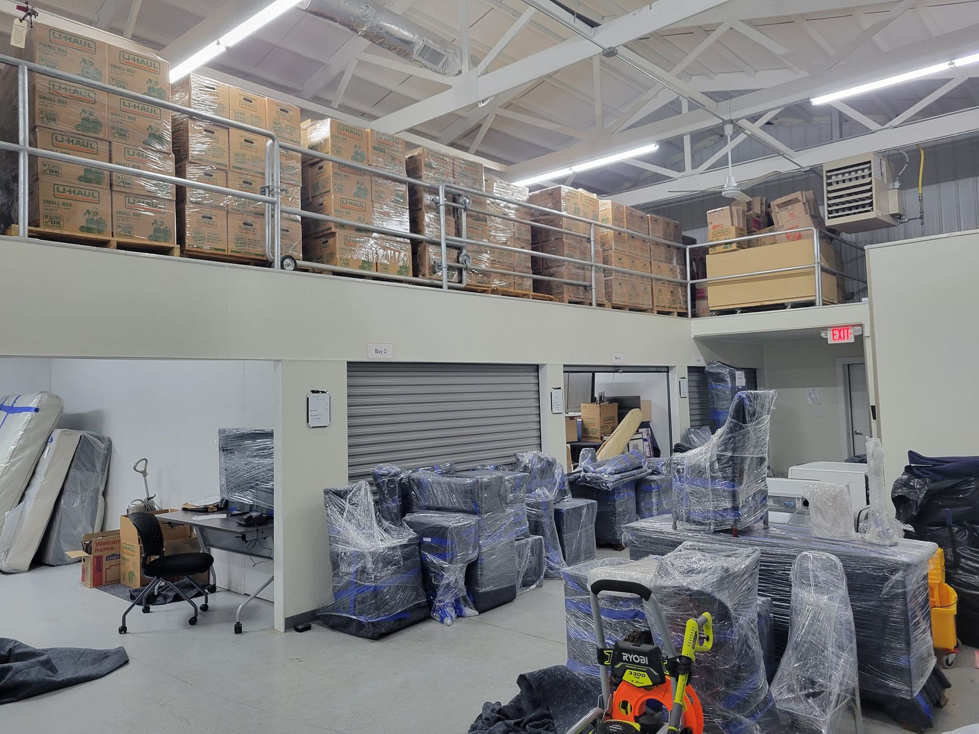 A warehouse interior featuring a mezzanine level stacked with cardboard boxes and a lower level filled with wrapped items.