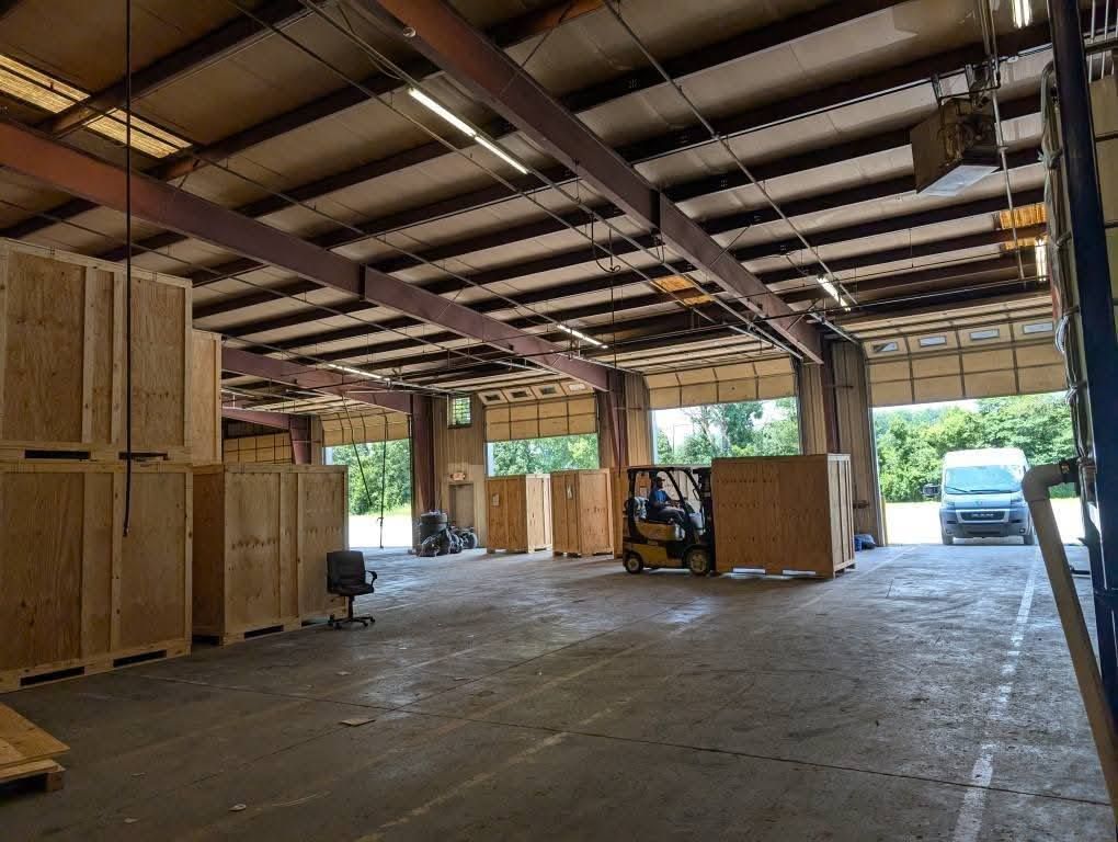 A forklift moves a wooden shipping crate inside a warehouse with tall ceilings and open bay doors.