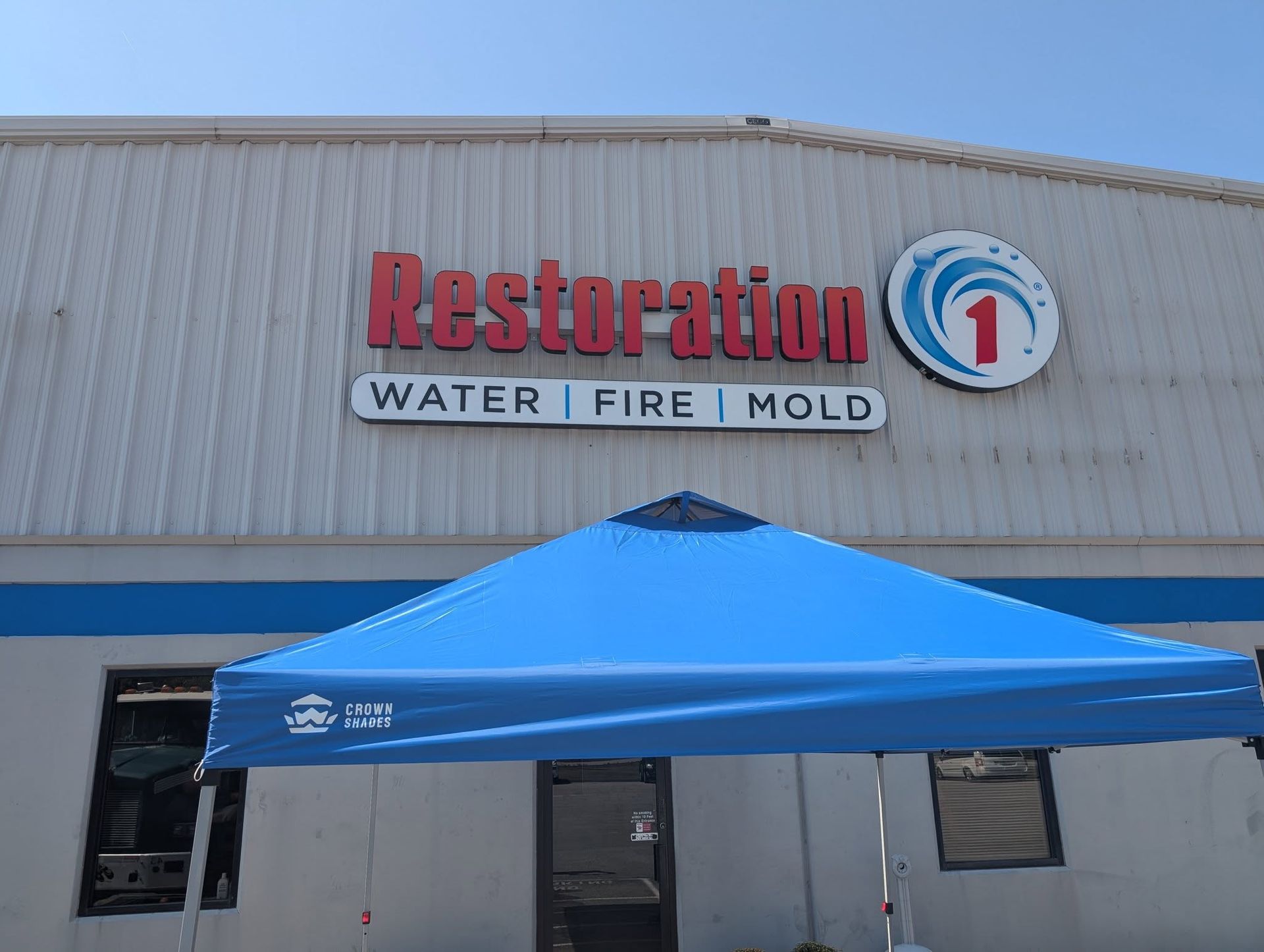 A blue pop-up tent stands in front of a Restoration 1 building sign, which reads