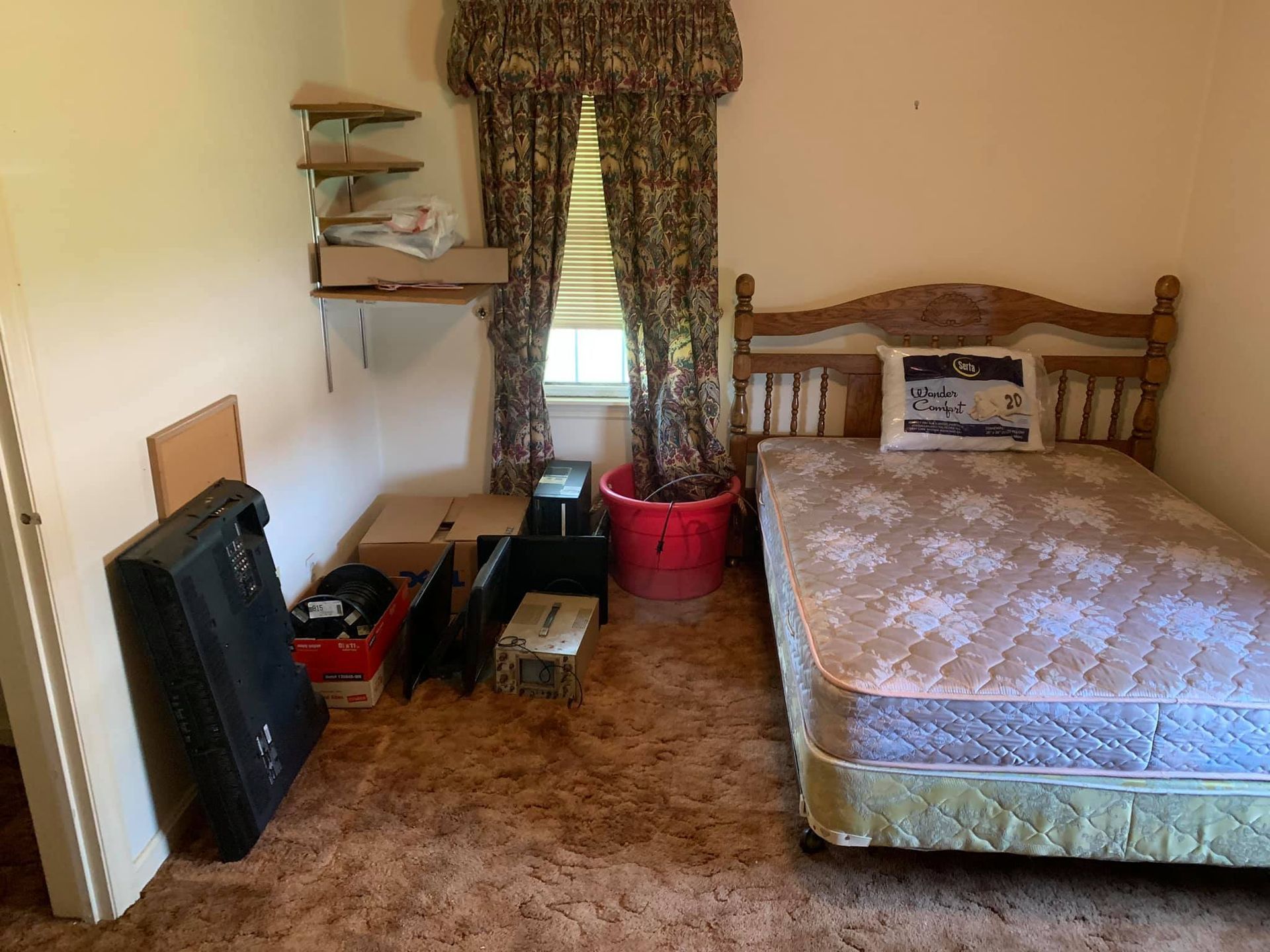 A bedroom with a wooden bed frame, mattress, floral curtains, stacked cardboard boxes, and a black flat-screen TV.