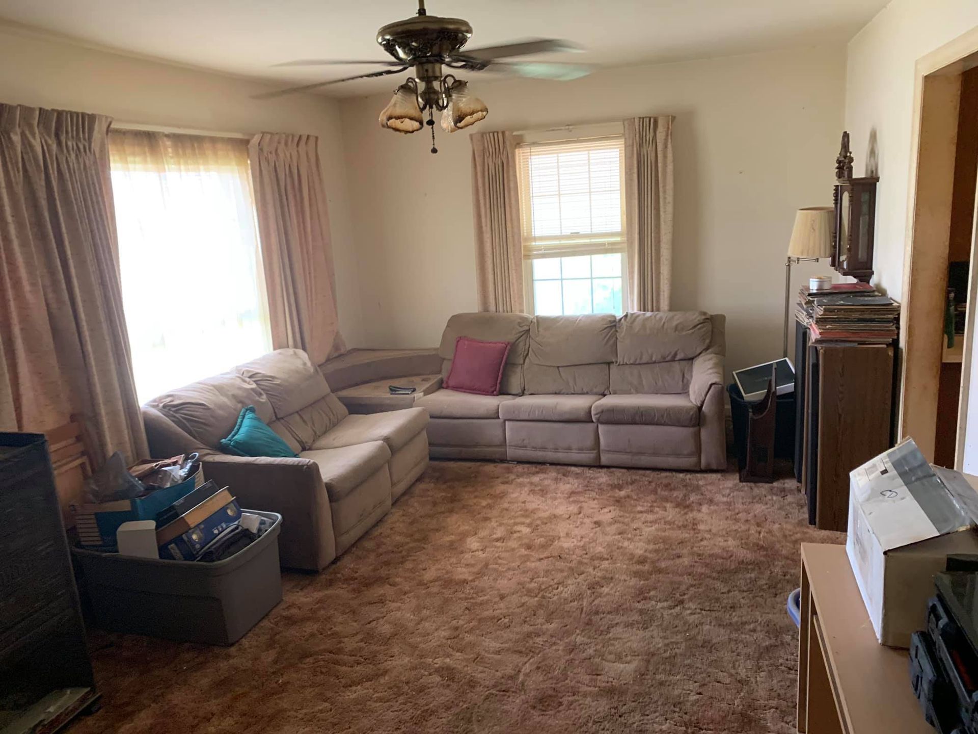 A carpeted living room with a large tan sectional sofa, beige curtains, a ceiling fan, and storage bins.