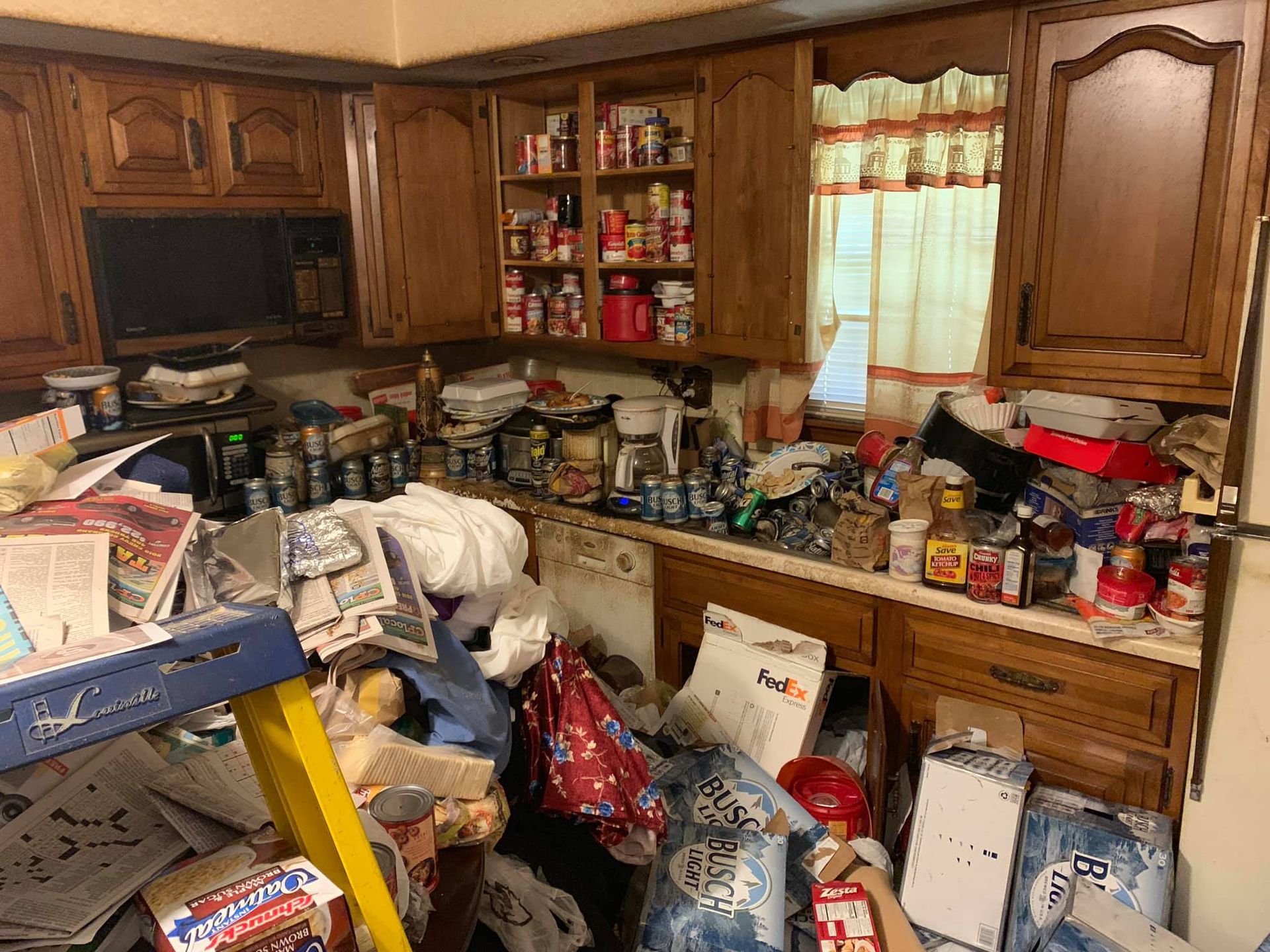 A cluttered kitchen featuring wooden cabinets, a window with curtains, and numerous items piled on counters and floor.
