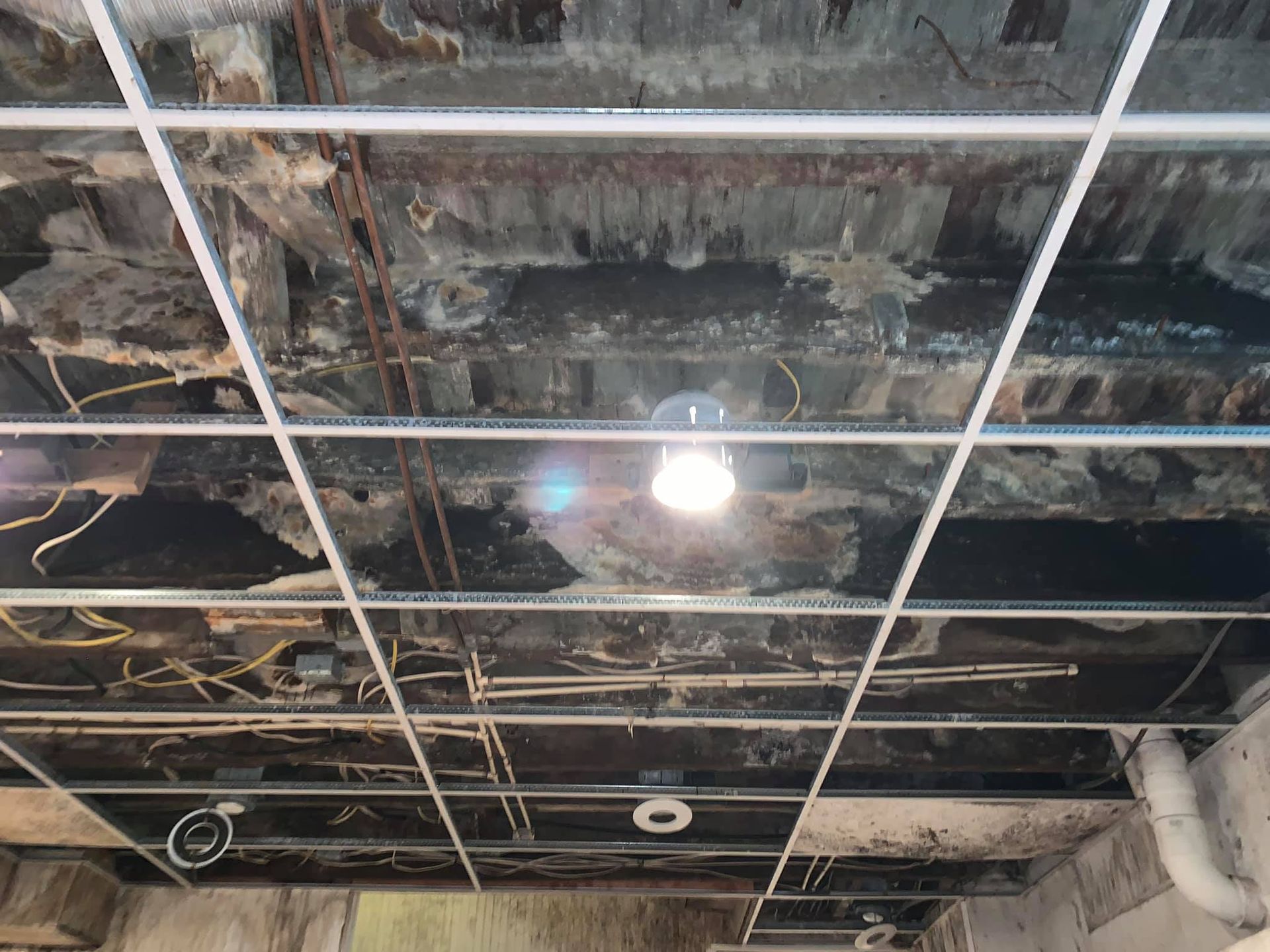 A view of a dark, unfinished ceiling structure with exposed beams, pipes, wires, and a metal grid frame.
