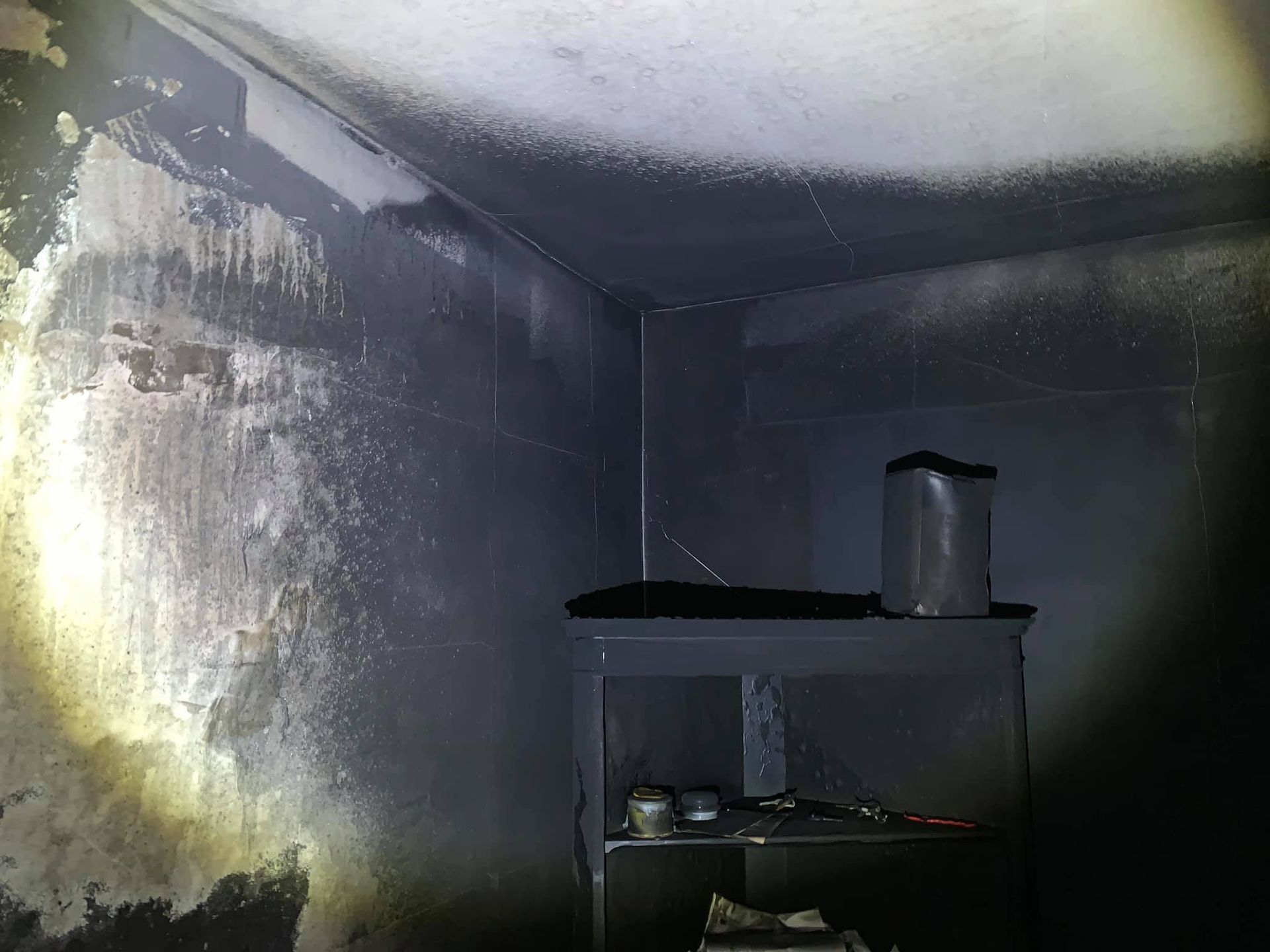 A dark, soot-covered room showing fire damage on the walls and ceiling, with a small table standing in the corner.