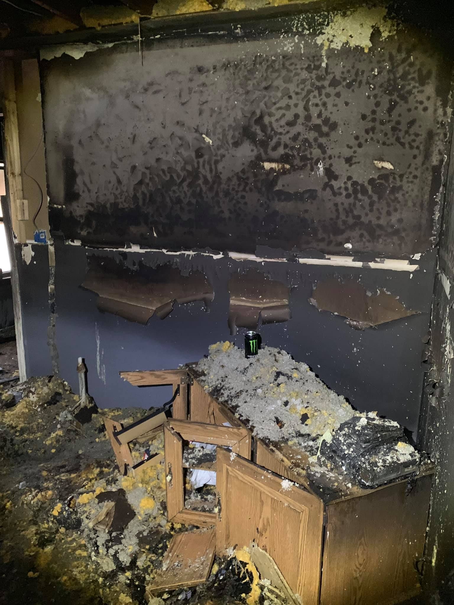 A charred room featuring a scorched wall, remnants of a wooden cabinet, and debris scattered across the floor.