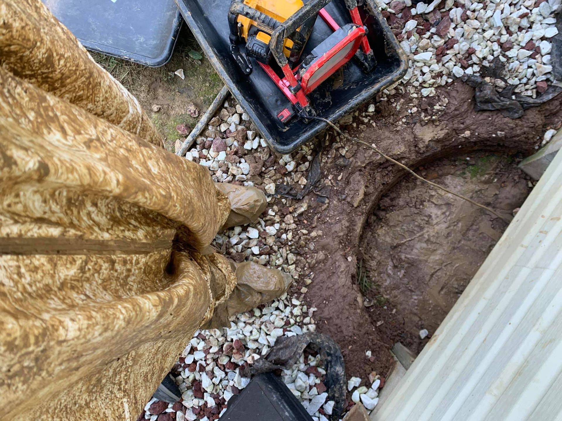 A person in camouflage clothing stands next to an excavated hole in the ground and a plastic bin containing a chainsaw.
