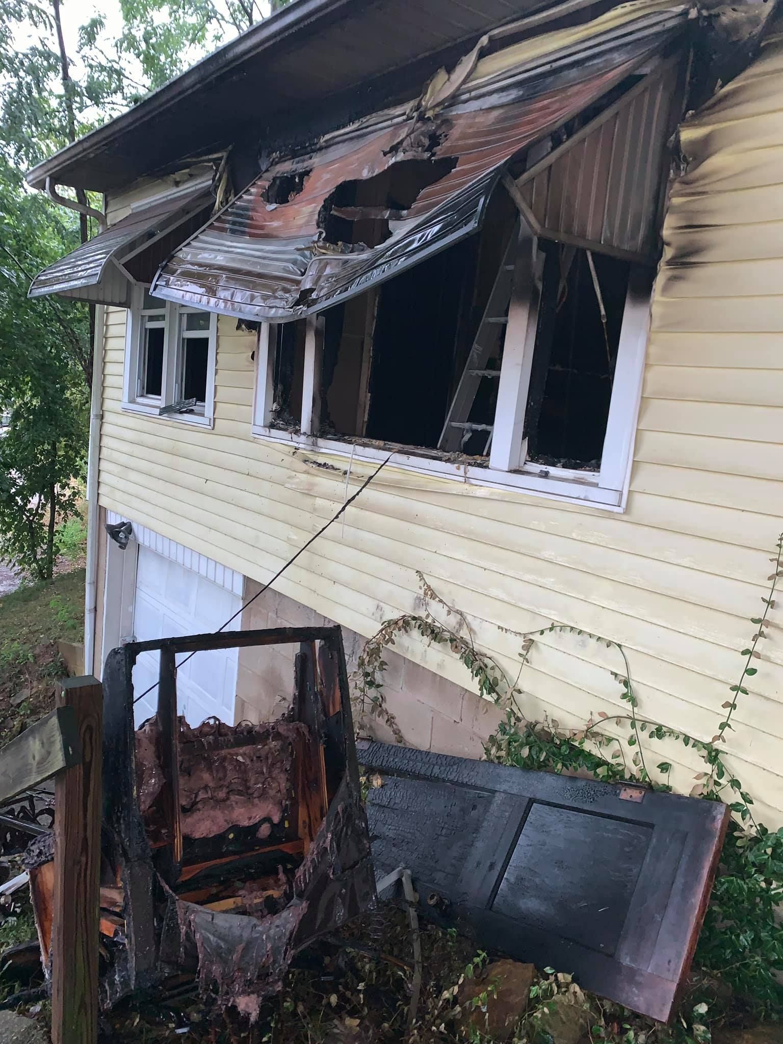 Fire-damaged yellow house with a charred exterior wall, broken windows, and debris on the ground below.