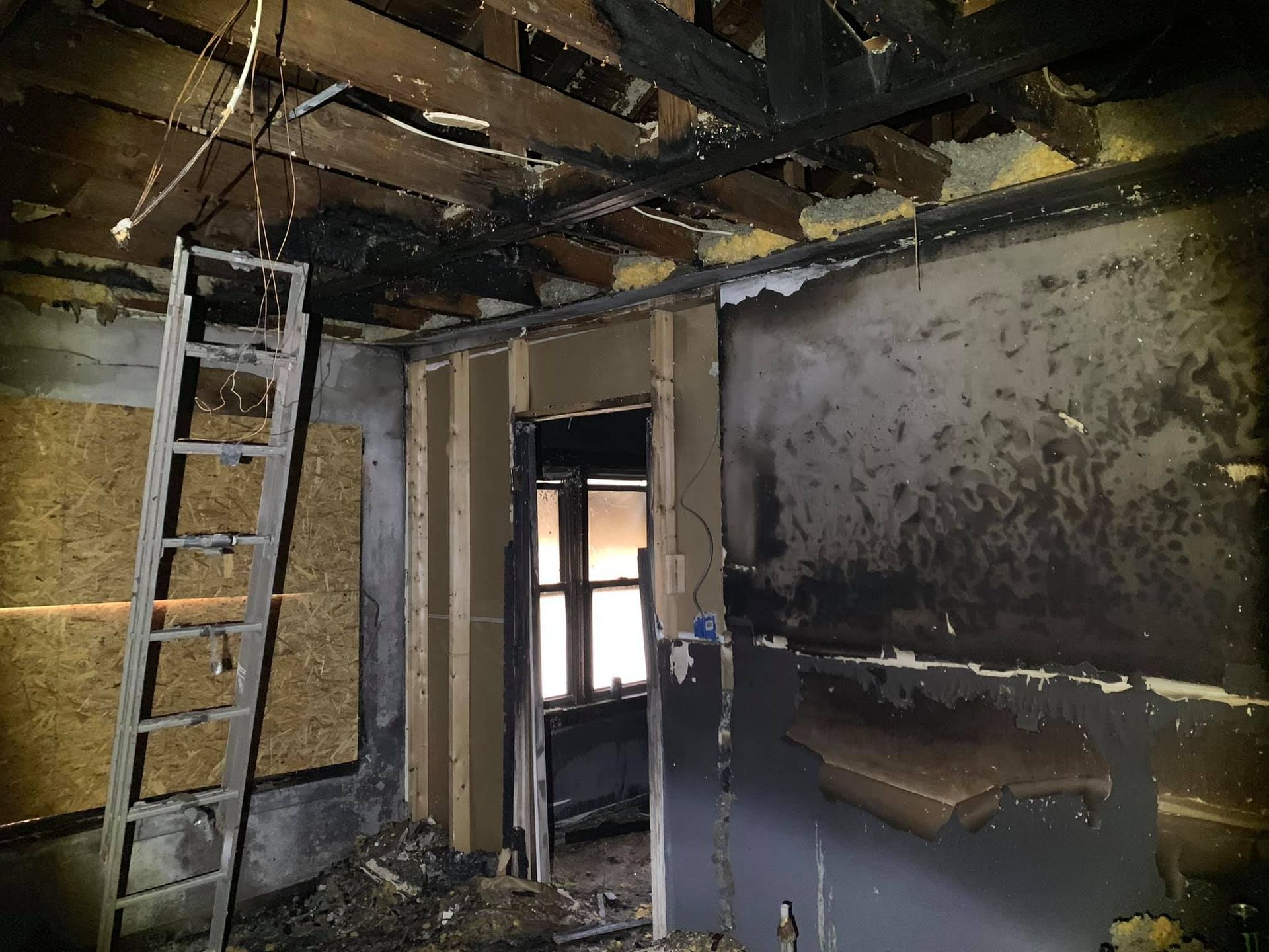 A room gutted by fire with scorched, black walls, exposed ceiling beams, and a metal ladder leaning against a wall.