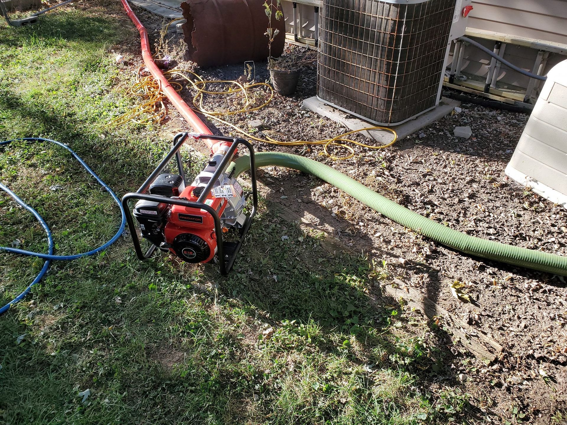 A portable gas-powered pump sits on grass with two hoses connected to it, located near a residential AC unit.