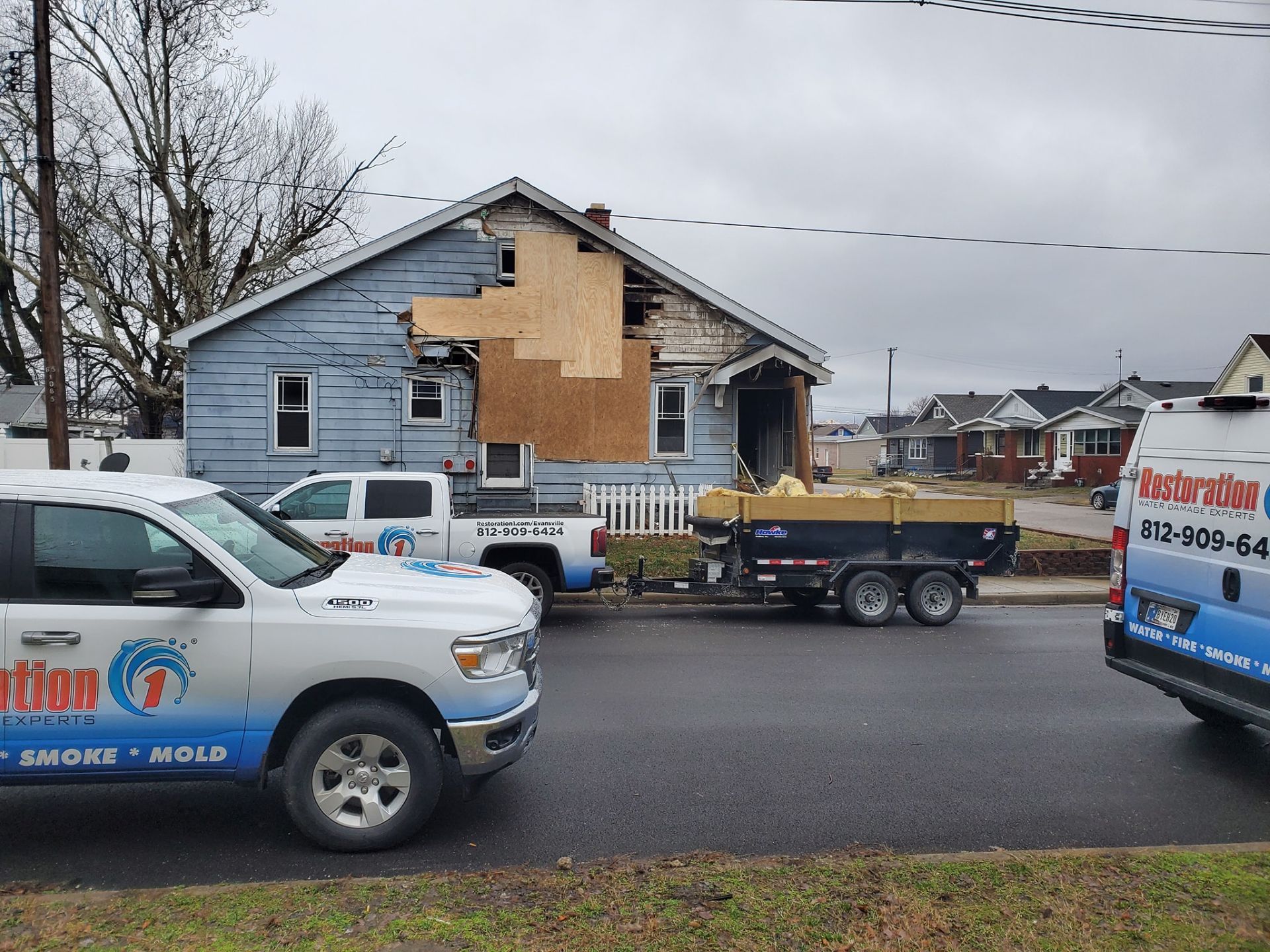 A damaged house with plywood boarding on the siding, flanked by restoration service vehicles and a utility trailer.
